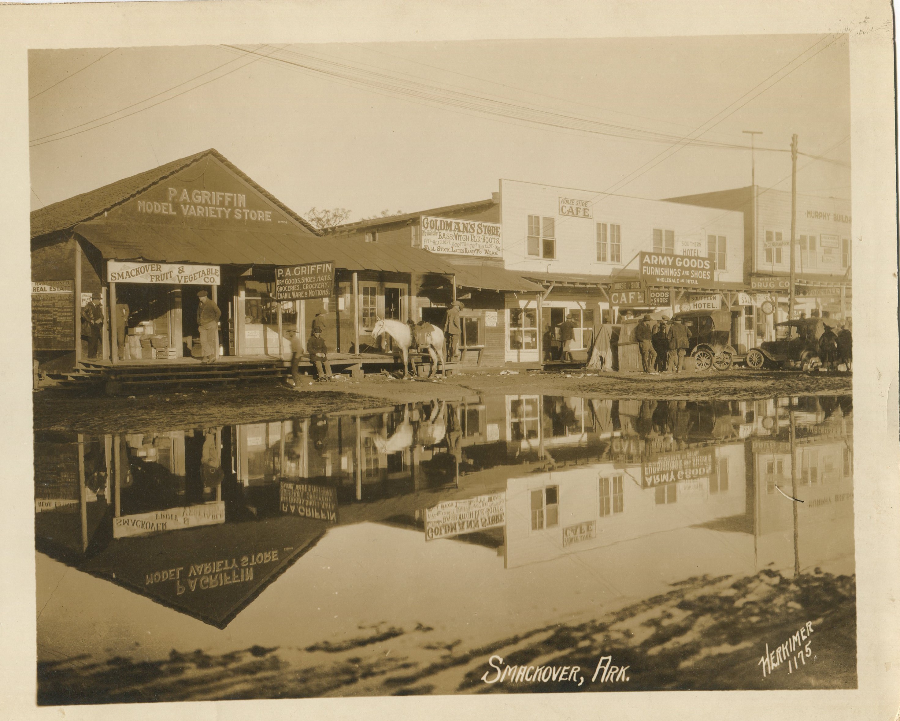 A daytime view of early Smackover. Wooden storefronts along a muddy road with a large puddle that has a mirror-like reflection of the storefronts. Cars, people, and horses are in front of the stores.
