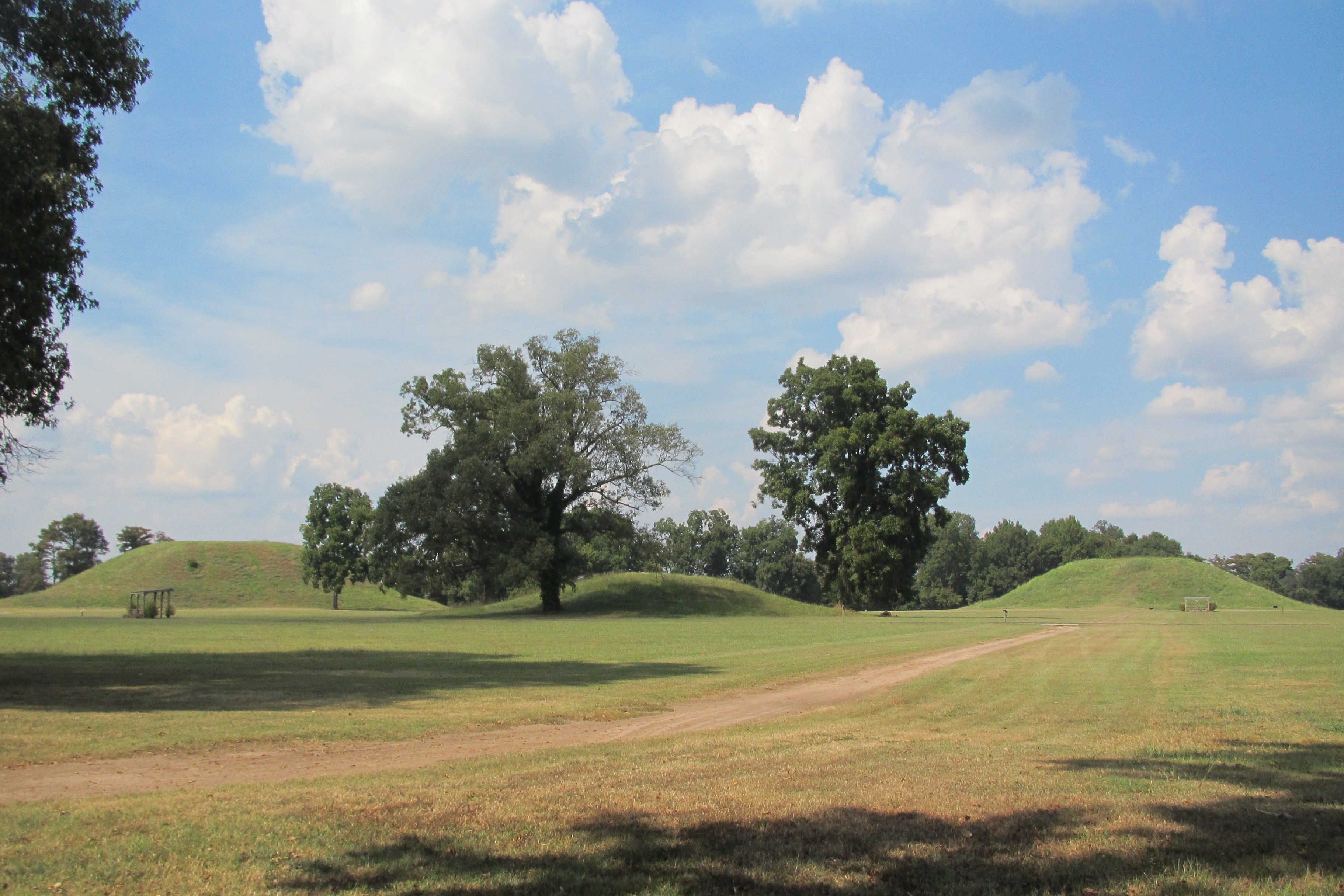 Three large mounds, Mounds A, C, and B, stand in an open grassy area with blue skies and puffy white clouds above. 