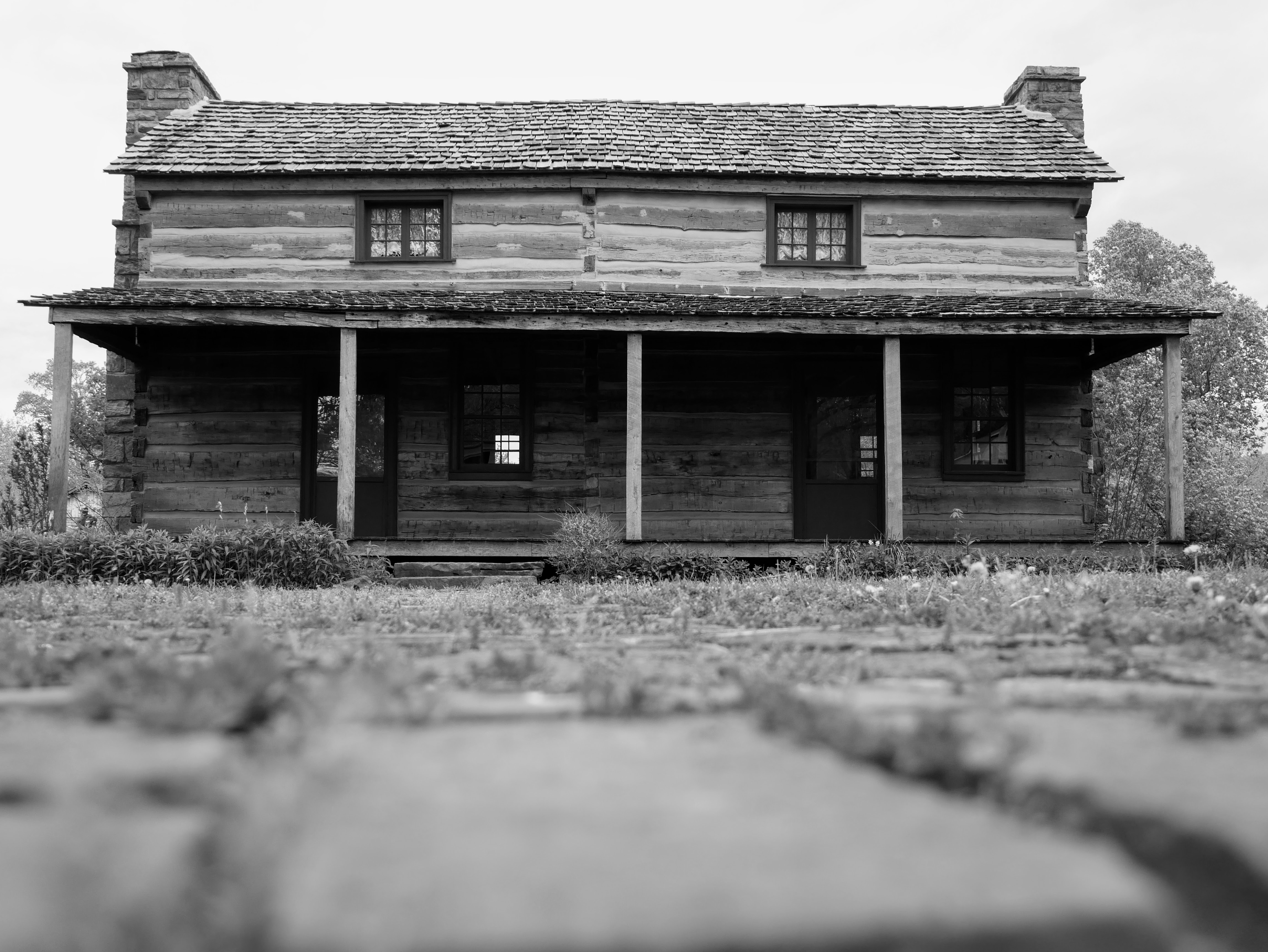 A two-story log home with large front porch. Two stone chimneys extend past the wood-shingled roof on each end.