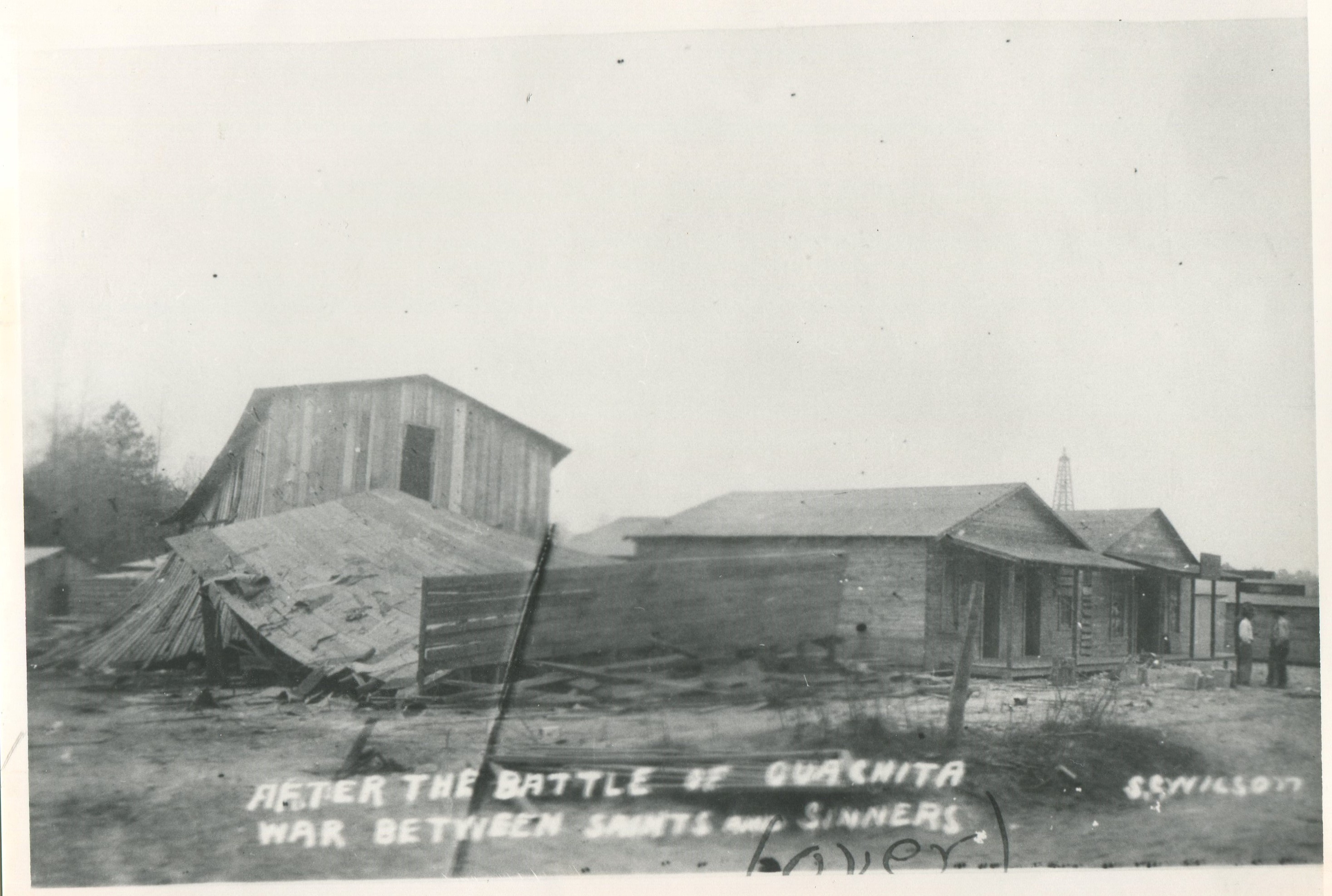 Broken wooden boards and other wooden debris of what is left from a barrelhouse scattered on the ground. Two wooden buildings to the right of the debris still standing. A view of an oil derrick is in the distance.  Writing on photograph says, “After the Battle of Ouachita War Between Saints and Sinners.”