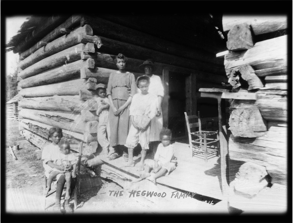 African American family of two women and five children posing for a photo on the porch of a log cabin home. A girl with a baby on her lap sitting on a chair in front of the porch. Two women and two boys standing on the porch and one child sitting on the porch. 