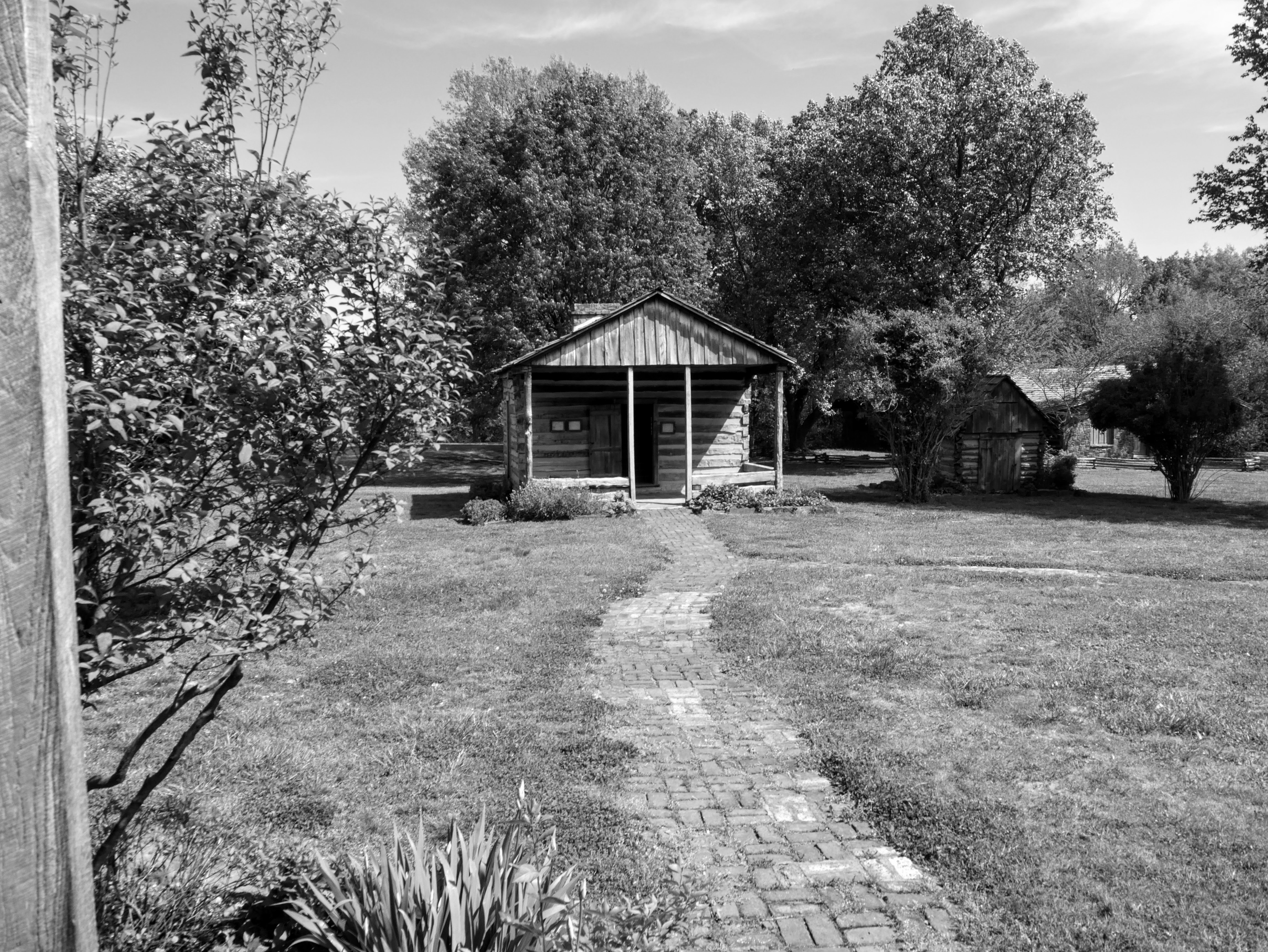 A meandering brick path leads to a small log building with large overhanging front-gabled porch.