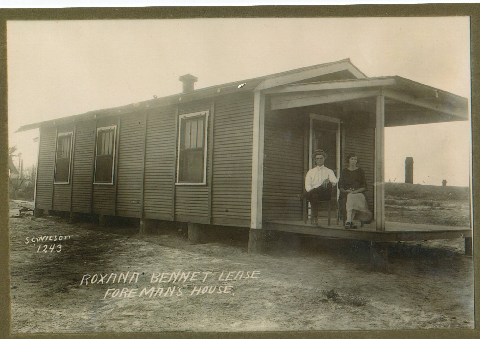 Well-dressed white couple sitting in chairs and posing for a photograph on the porch of a wooden shotgun house. Man is wearing a hat, white shirt, and tie with dark-colored pants, woman is wearing a dark-colored blouse with a light-colored skirt. The house is a dark color with white trim on the front. 