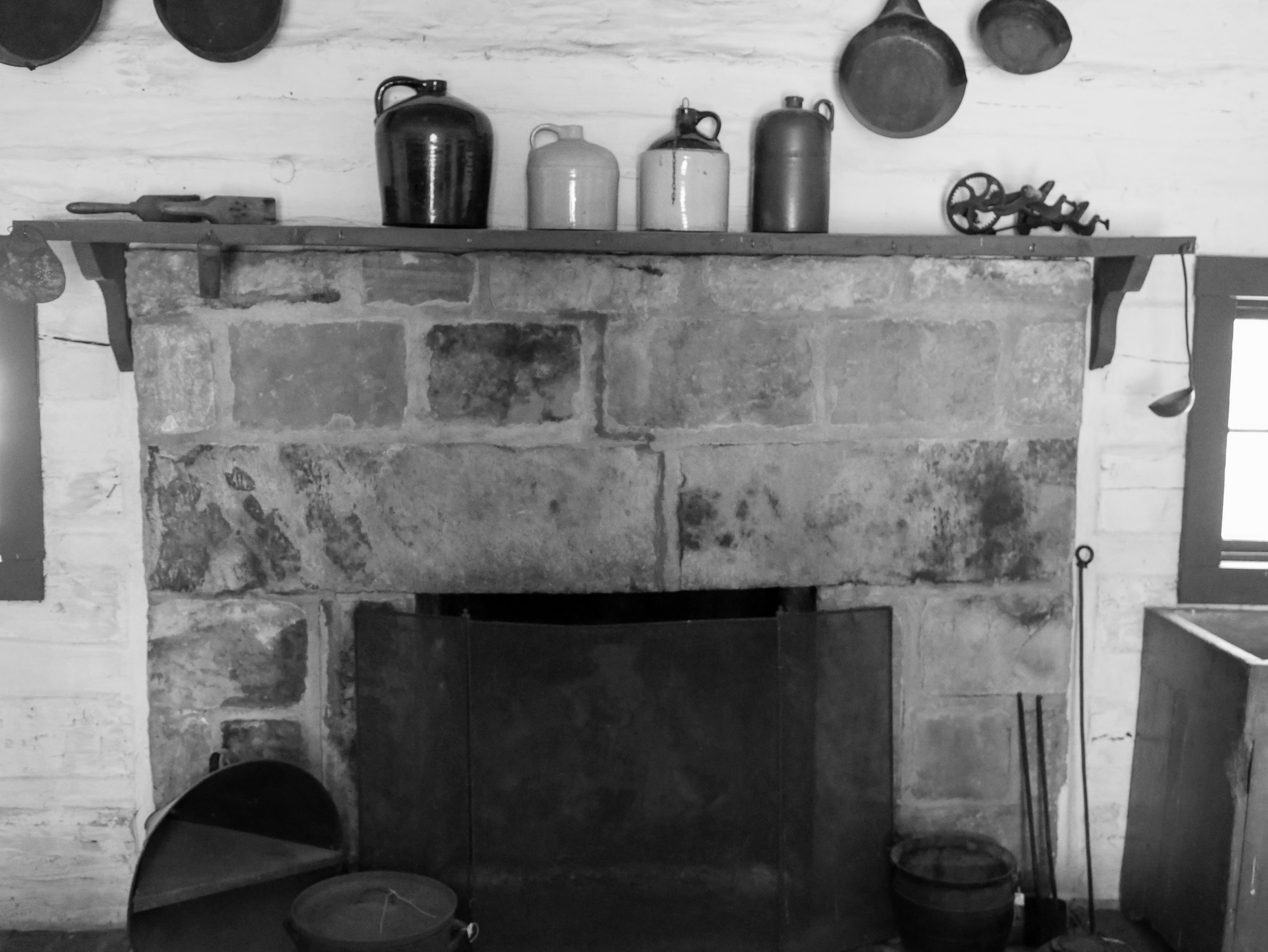A large stone fireplace with iron screen is in the middle of the back wall of the kitchen. Several large iron Dutch ovens and fireplace utensils are set at each side. Atop the wooden mantle sit several large ceramic jugs. Iron skillets are hung barely out of frame above the fireplace.