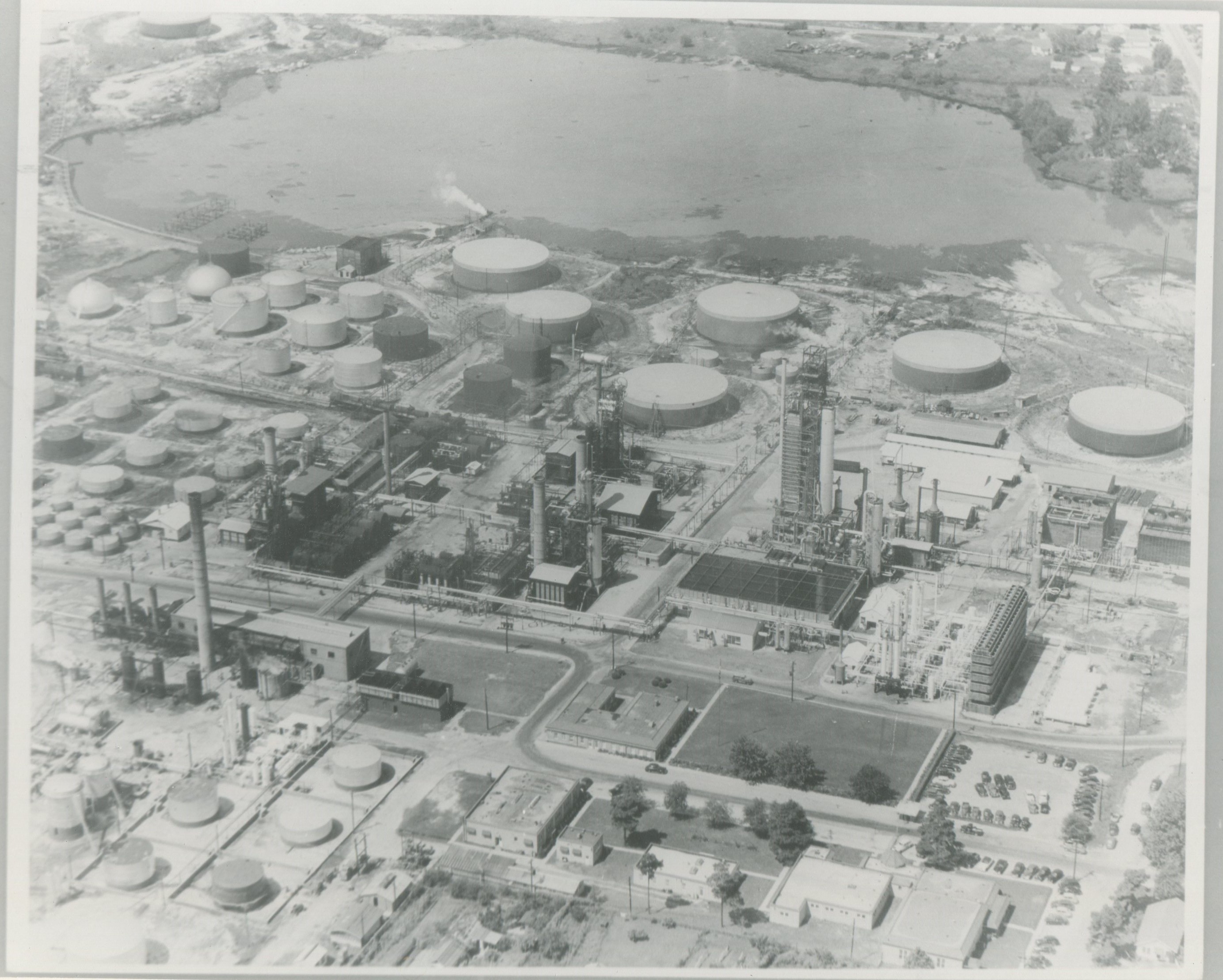 Aerial view of a working oil refinery with scattered buildings, smokestacks, pipelines, oil storage tanks, a large body of water and cars in a parking lot. 
