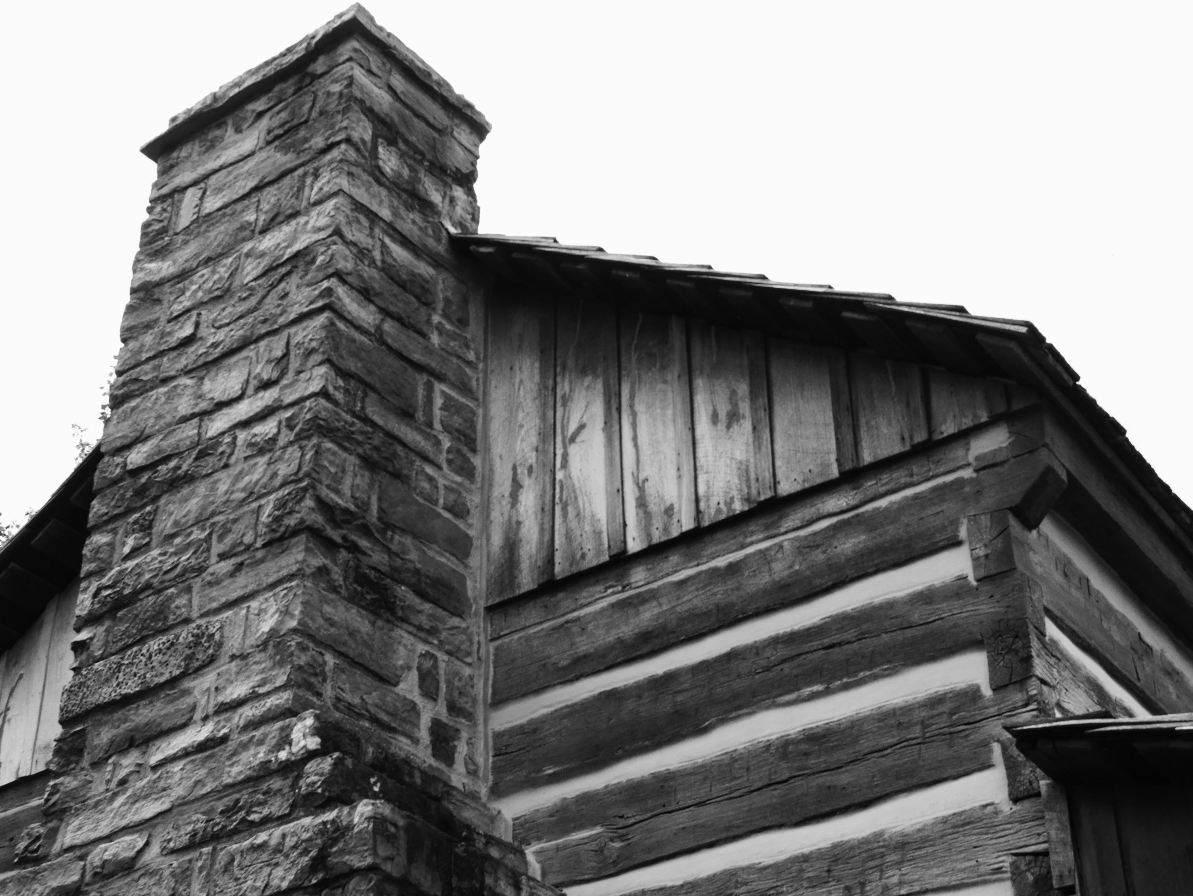A chimney made of rectangular stones extends past walls made of square logs with half-dovetail notching and gable covered in vertical board and batten wooden siding. Overlapping wooden shakes are barely visible at the edges of the roof.