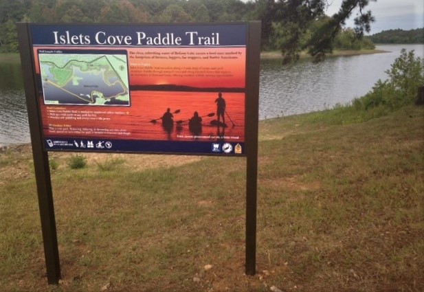 view of the Islet Cove Trailhead sign with lake and kayak put-in area in background 