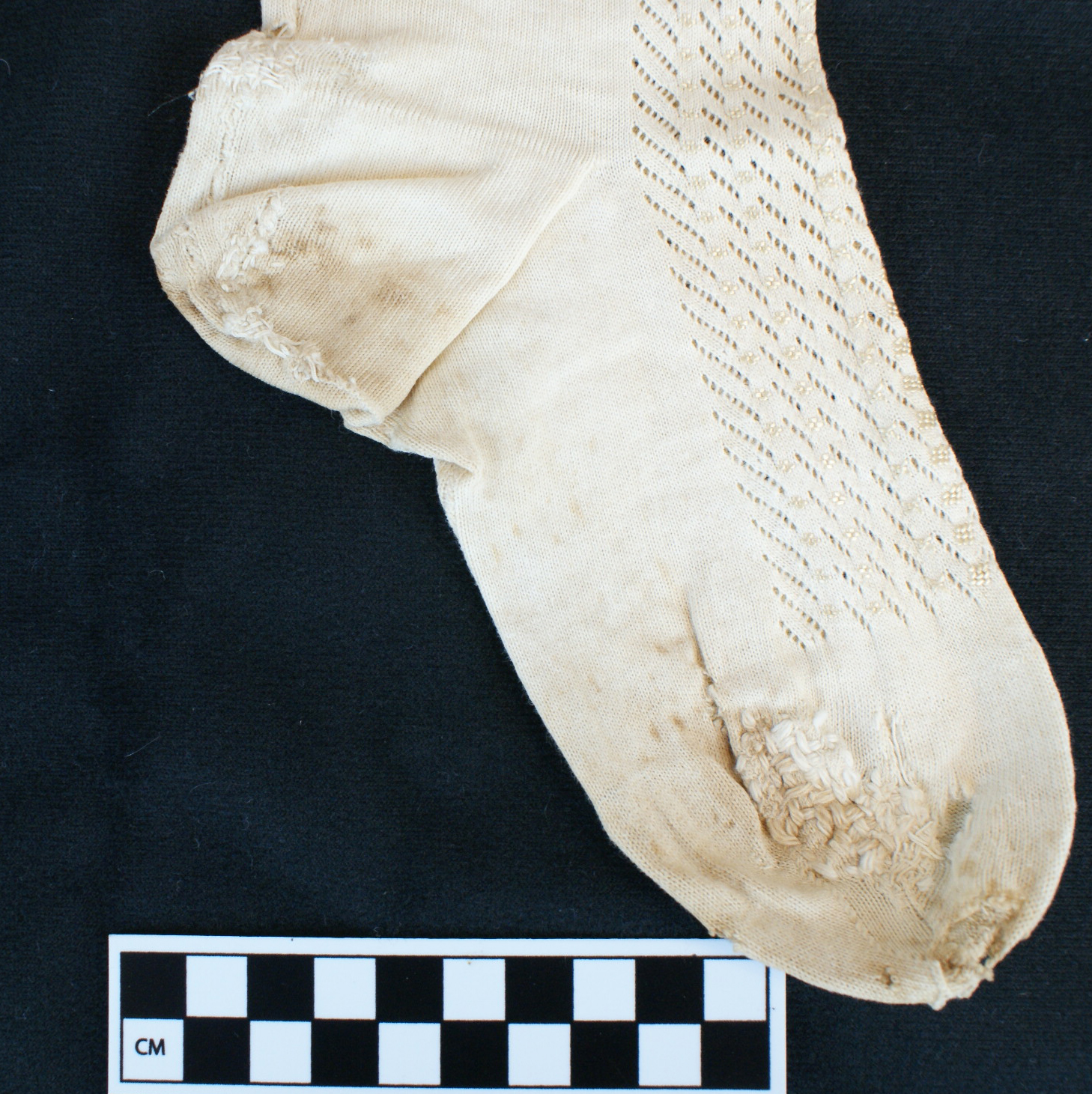 detail view of the foot of a white stocking against a dark background, with a centimeter scale in the lower left so the viewer can gauge the artifact’s size.  