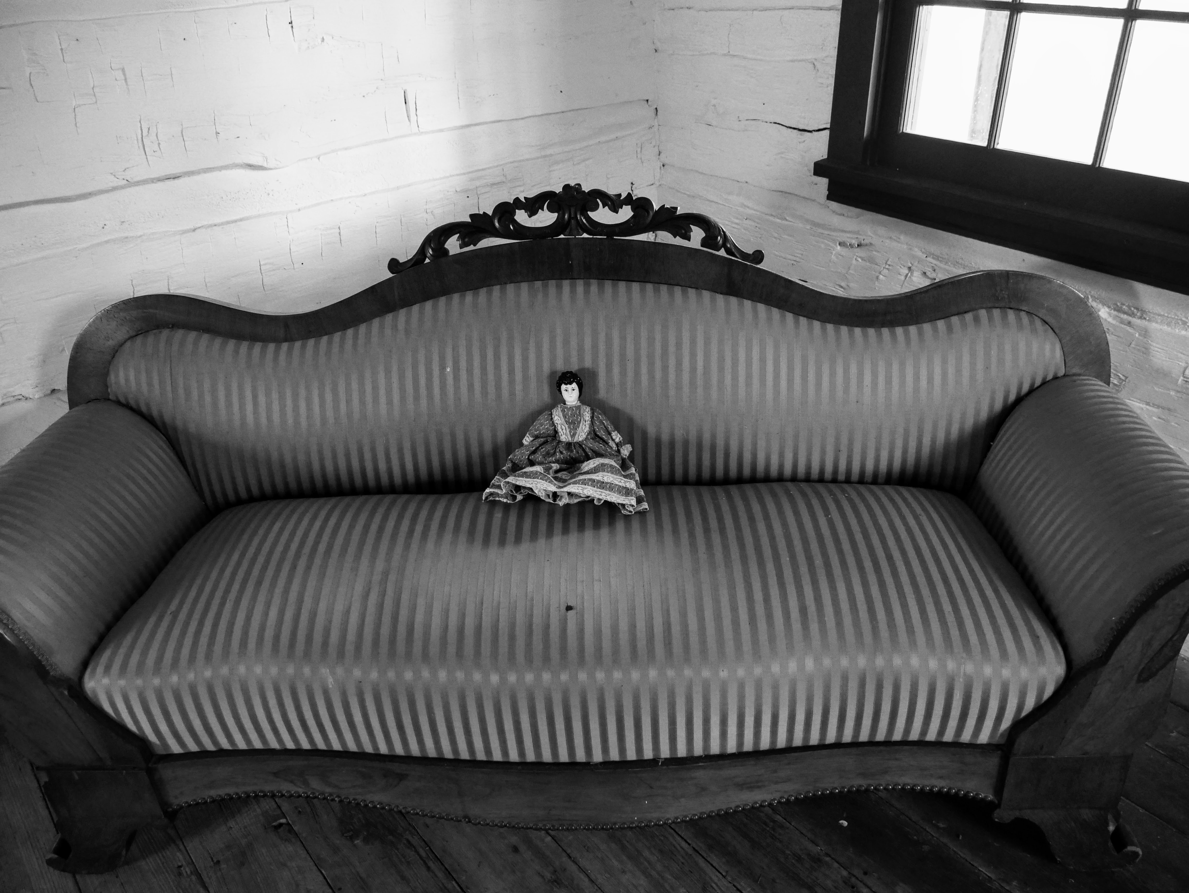 In the corner of a white-washed room, a wooden couch with striped upholstery is sat at an angle. A small ceramic doll in a fabric dress is in the middle of the couch.