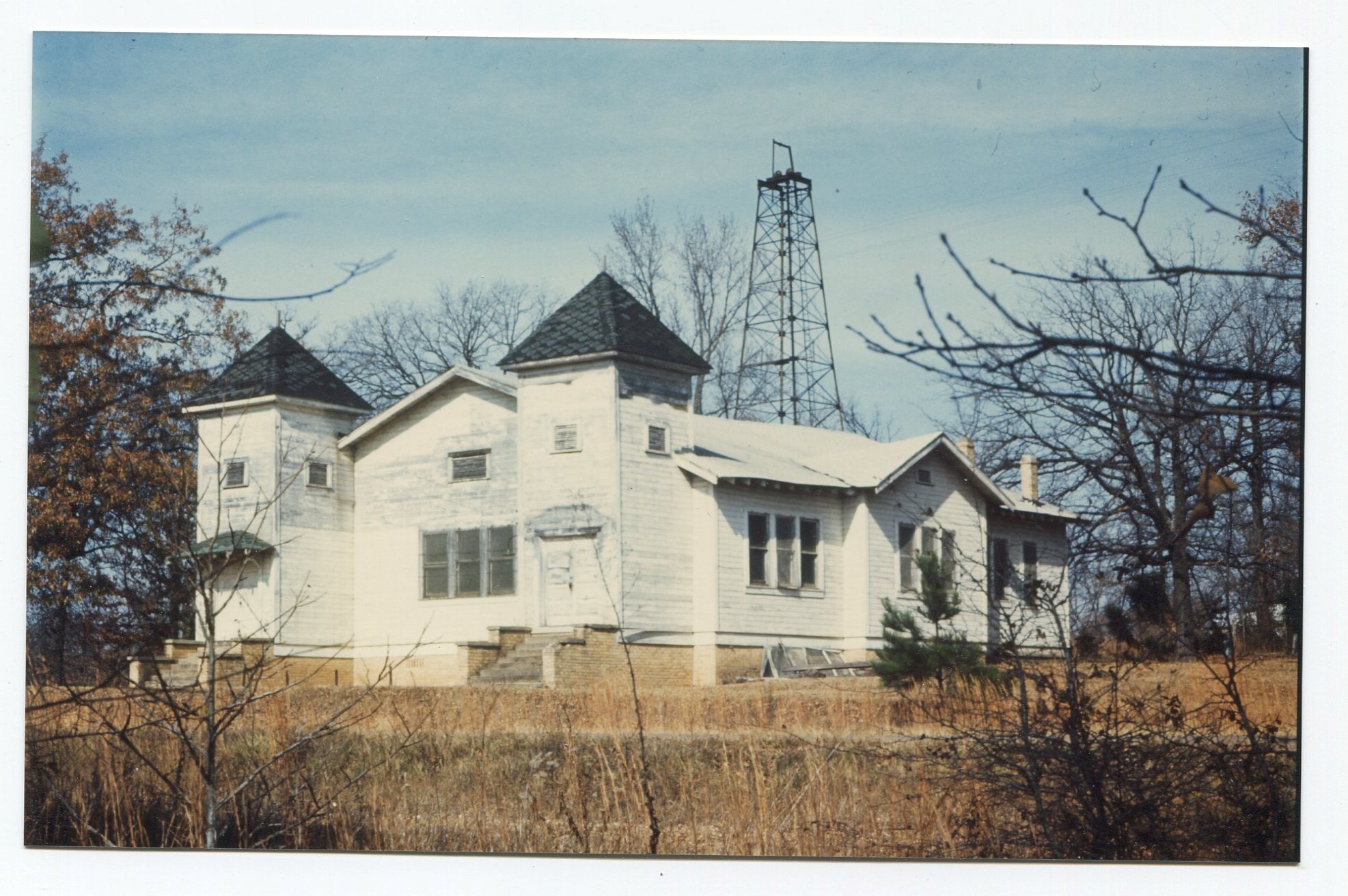 A ground-level view of a large, white wooden church and the surrounding area covered with brown grass and trees with a metal oil derrick in the distance behind the church.