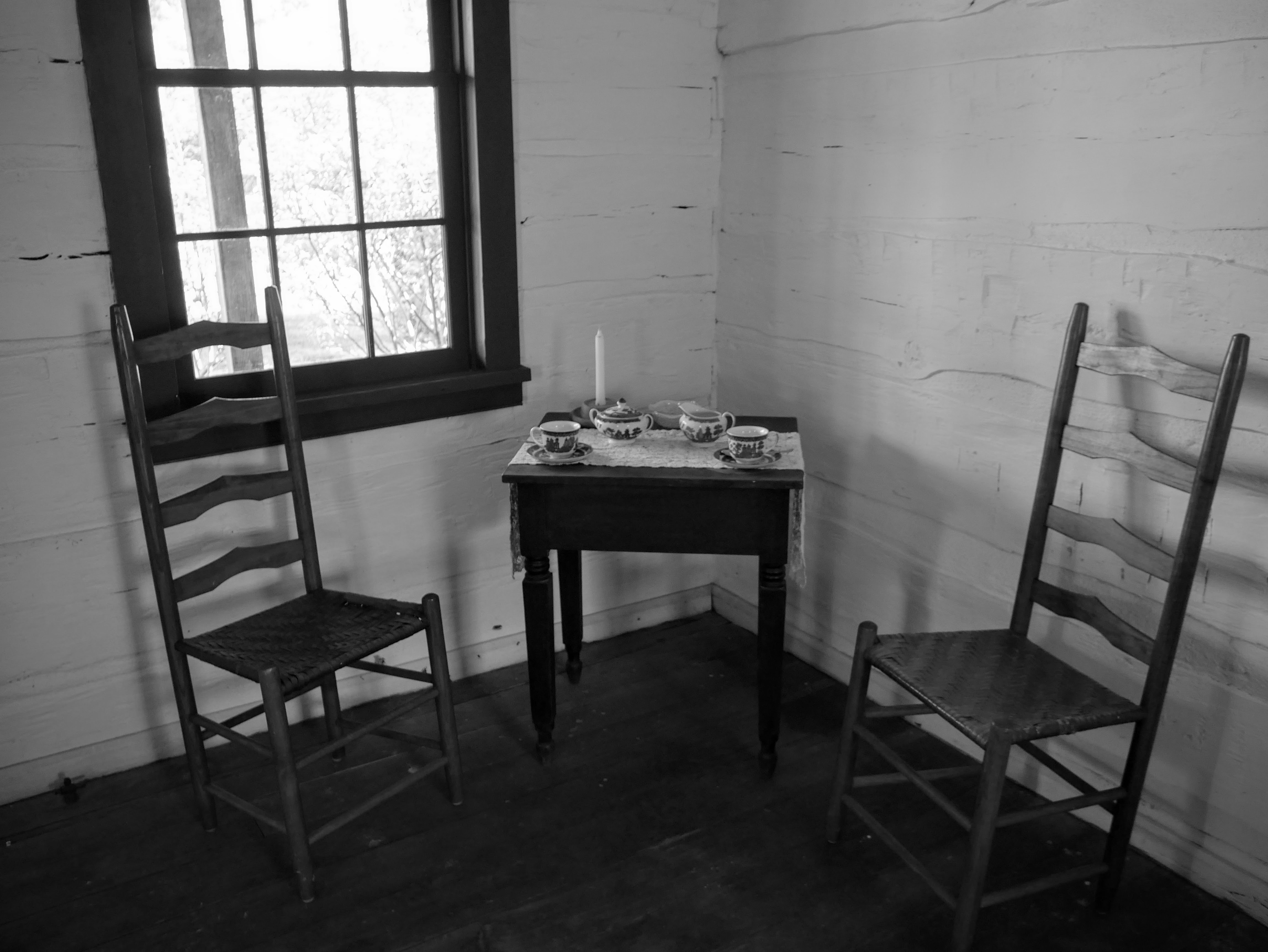 In the corner of a whitewashed room, two straight-backed chairs flank a small side table. Atop the table are two teacups, a sugar bowl, creamer, and one candle.