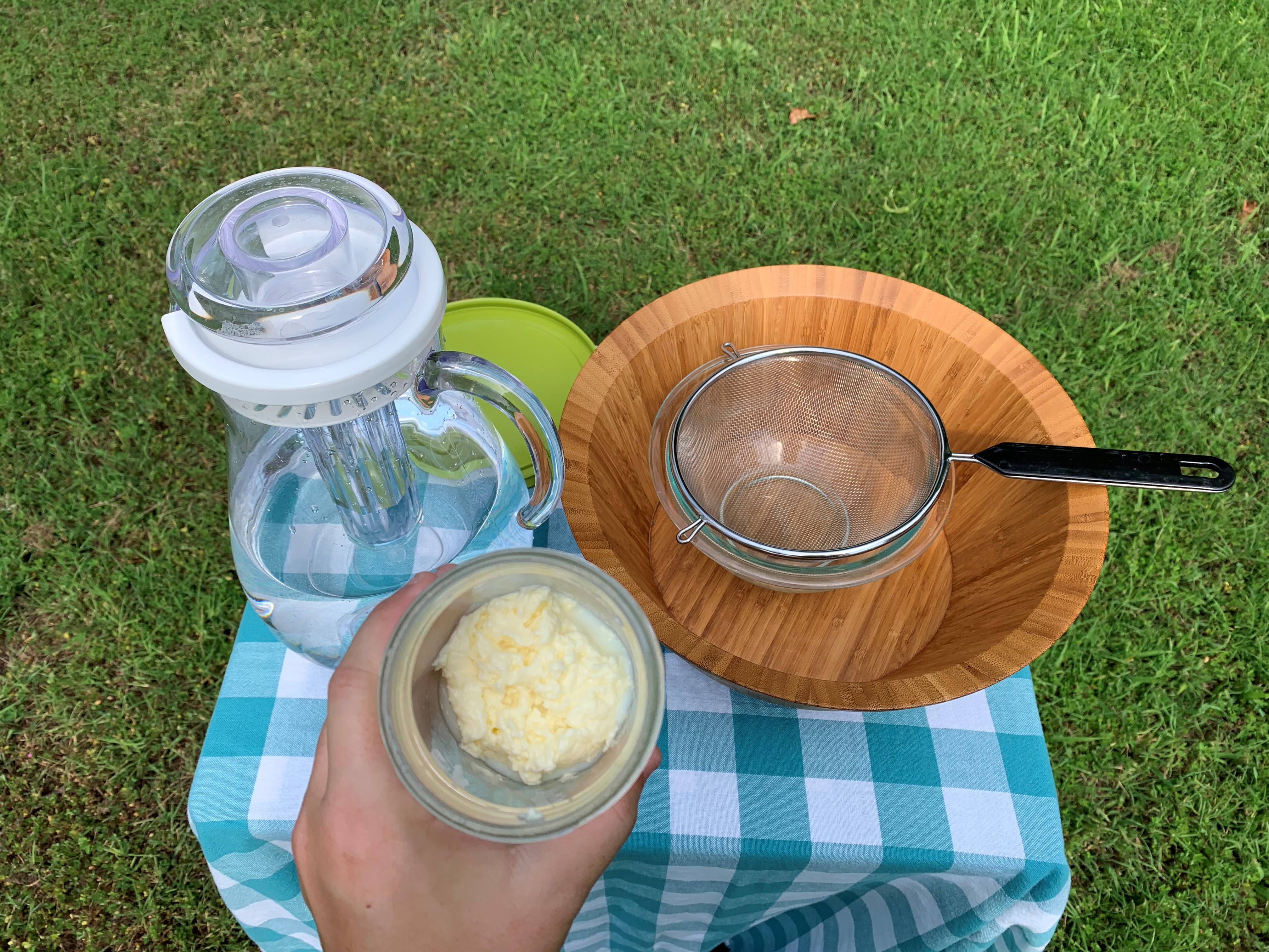 a hand holds a mason jar to show the ball of butter that has formed inside, on the table is materials for the next step: two bowls a strainer and a pitcher of water