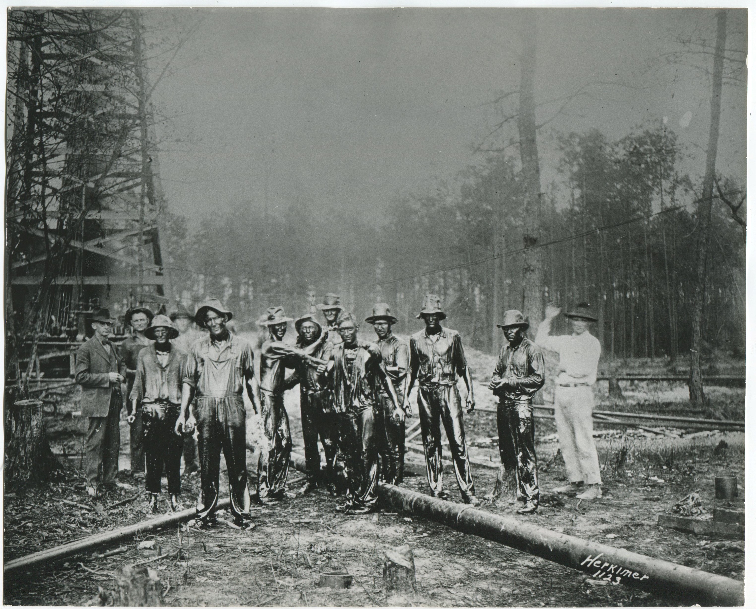 Group of oil field workers dressed in work clothes and covered in oil stand in front of a wooded oil derrick and pose for a photo with a man on the left side of the group dressed in a suit not covered in oil and a man on the right dressed in white and waving.  