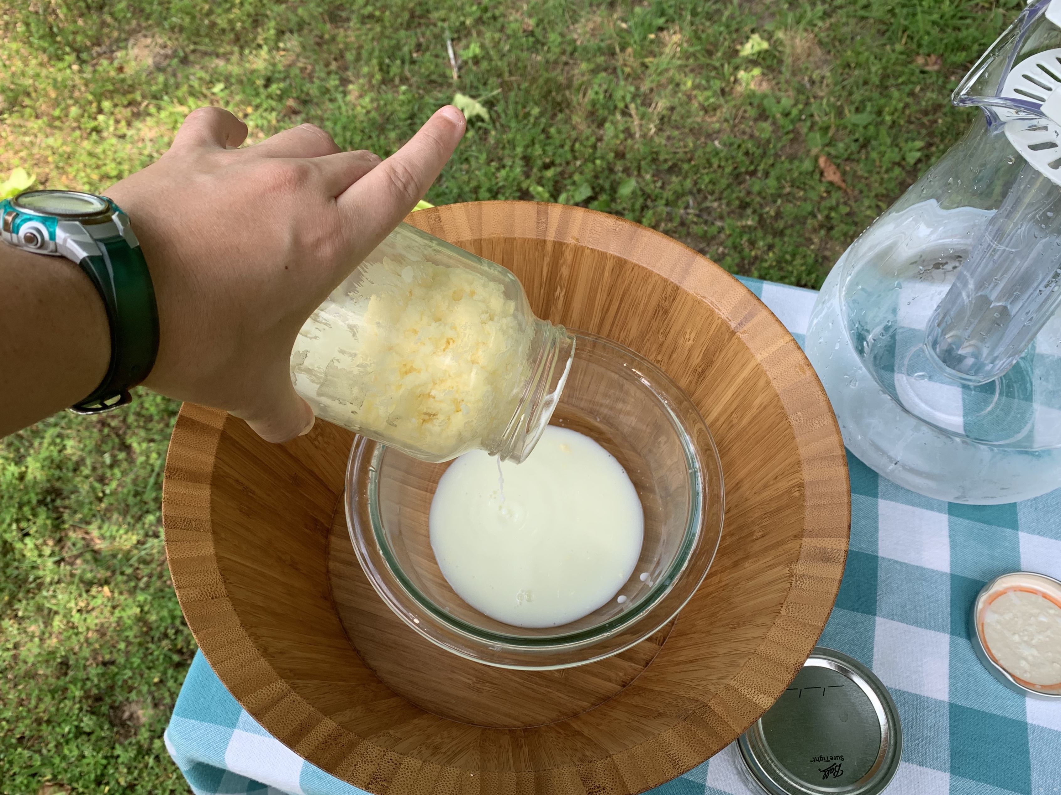 A hand is holding a mason jar with a ball of butter in it, on the table there are two bowls one is for catching the buttermilk and one is for the next step. The buttermilk is being poured out of the jar into one of the bowls