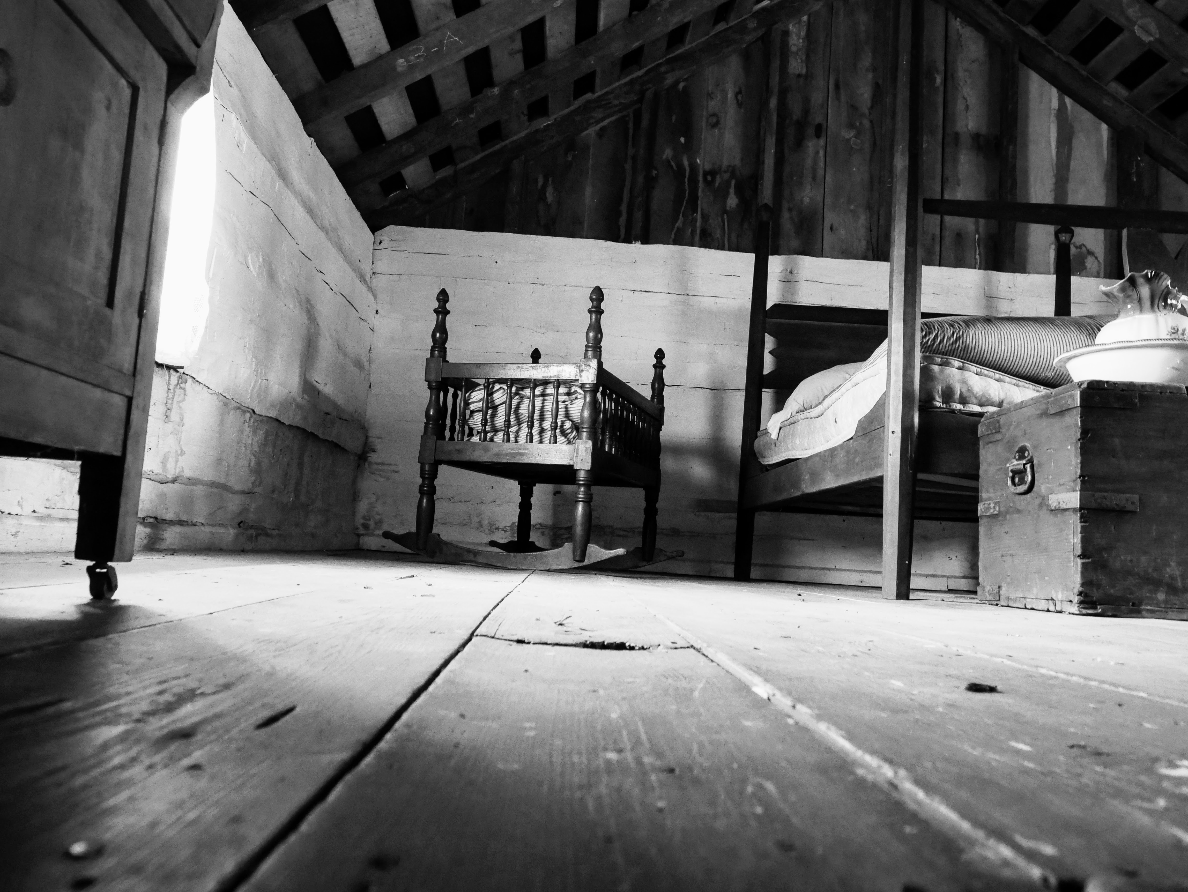The image is taken looking up from the floor and features rough, uneven floorboards in the foreground. At the back wall of the whitewashed room, a large four poster bed and wooden trunk are sat to the right of a small, rocking bassinet. The gable end of the room and the roof are unfinished.
