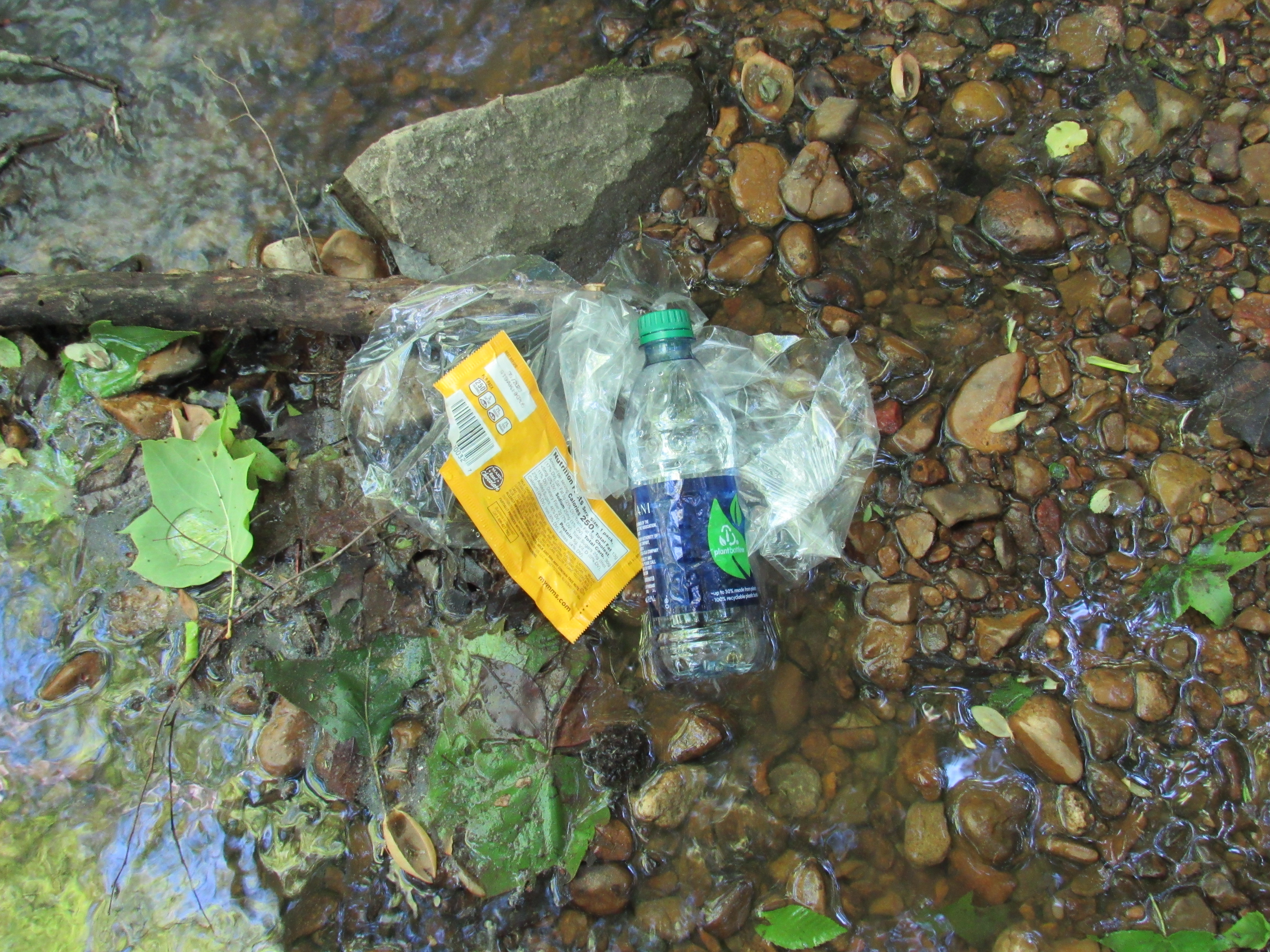 A plastic water bottle, candy wrapper, and plastic bag laying in a creek 