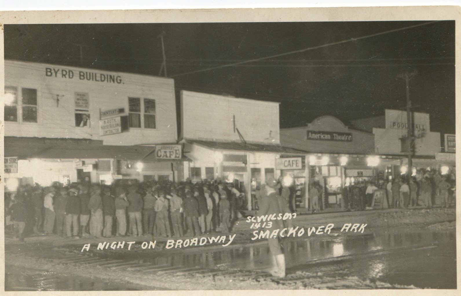 A night view of large crowds of people standing in front of wooden store fronts in Smackover. Buildings include two cafes, a theater, and a pool hall. A large water puddle in the road in front of the stores and a man is crossing on wooden planks over the mud. Writing on the photograph says, “A night on Broadway.”