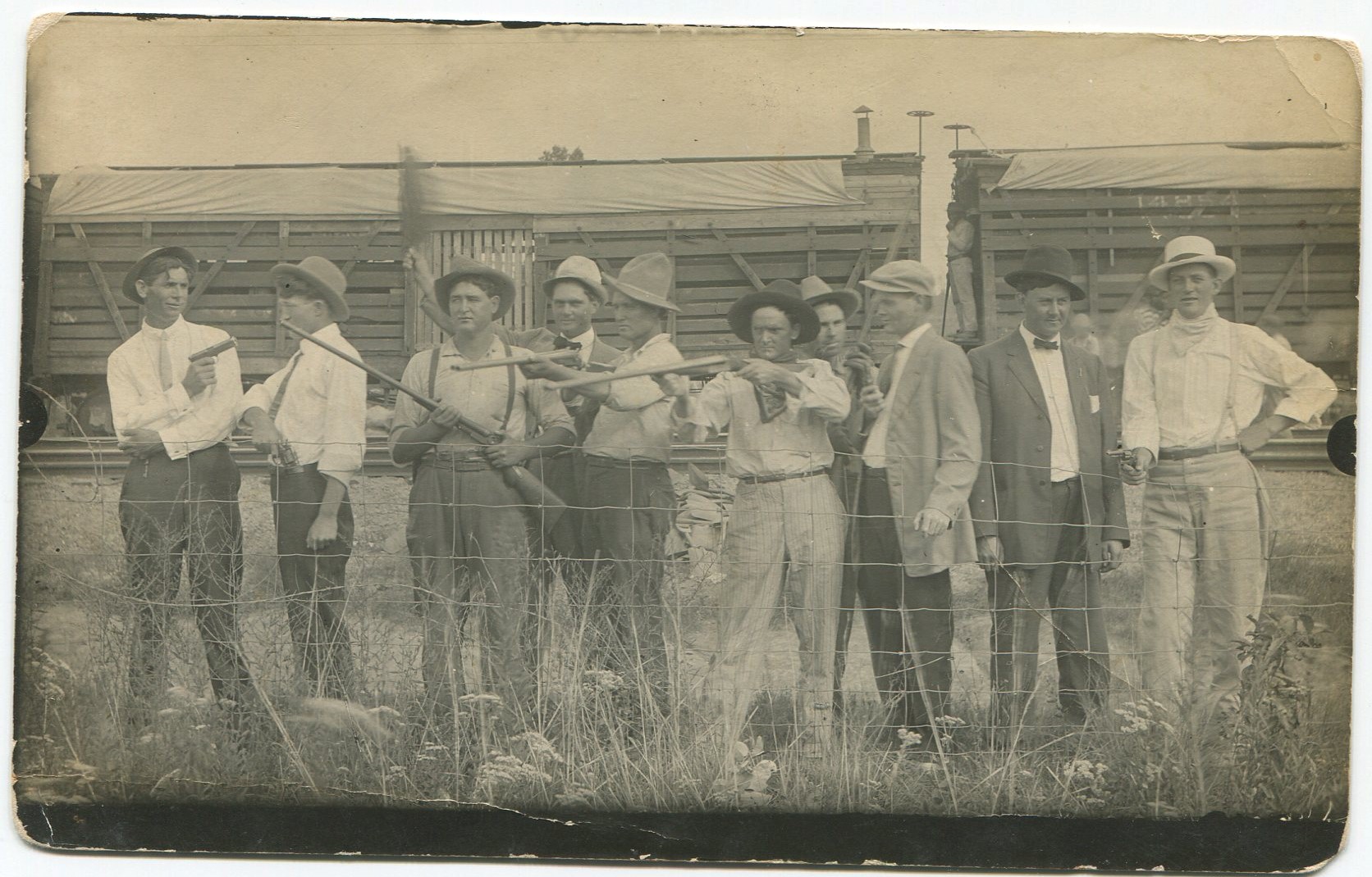A group of men holding and pointing rifles and handguns, while standing behind a metal wire fence and in front of wooden railroad boxcars with a few onlookers scattered about.   