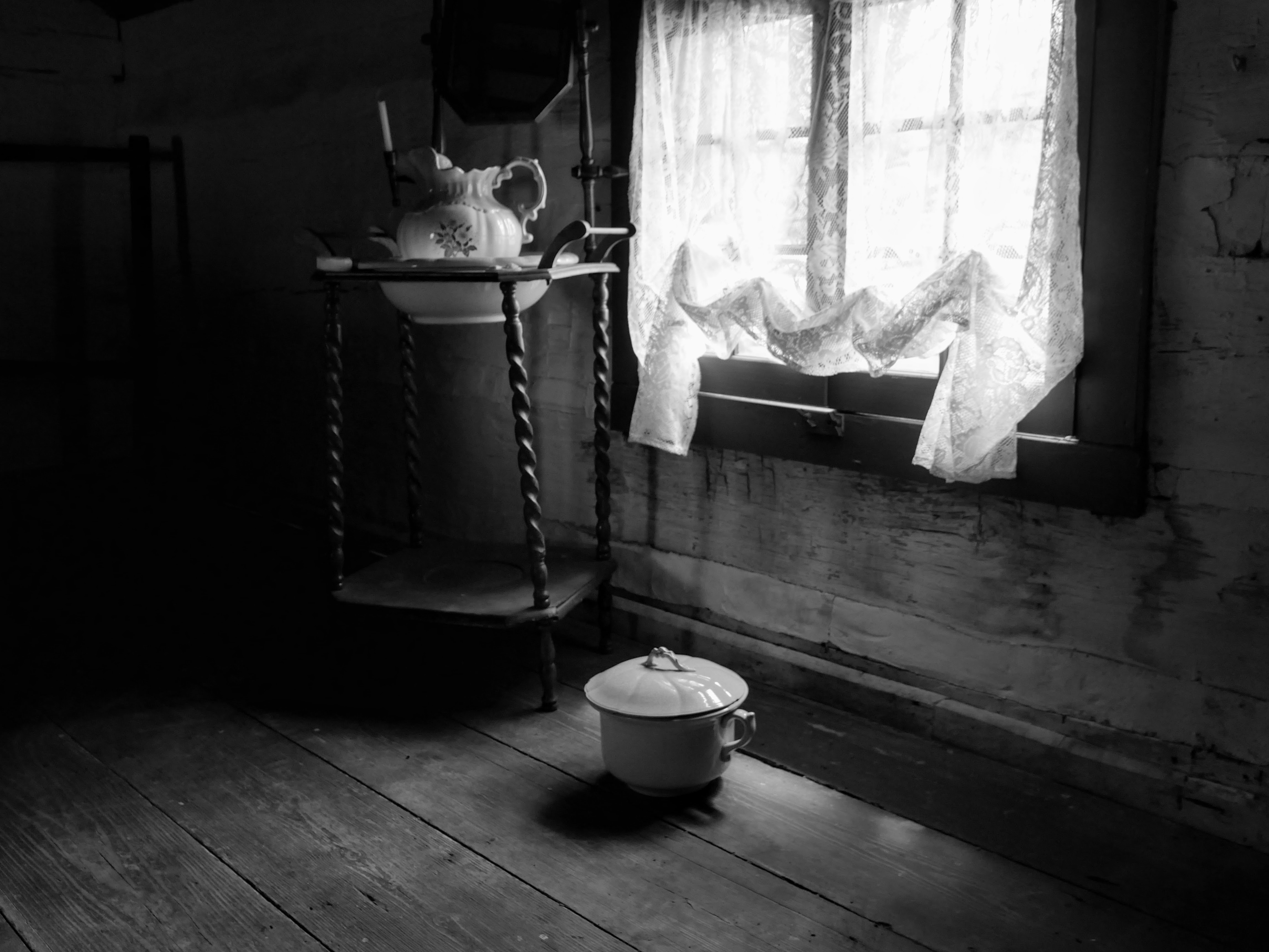 A small window with lace curtains illuminates a wooden washstand with mirror, bowl, and pitcher. On the wooden floor underneath the window sits a white, ceramic chamber pot with lid.