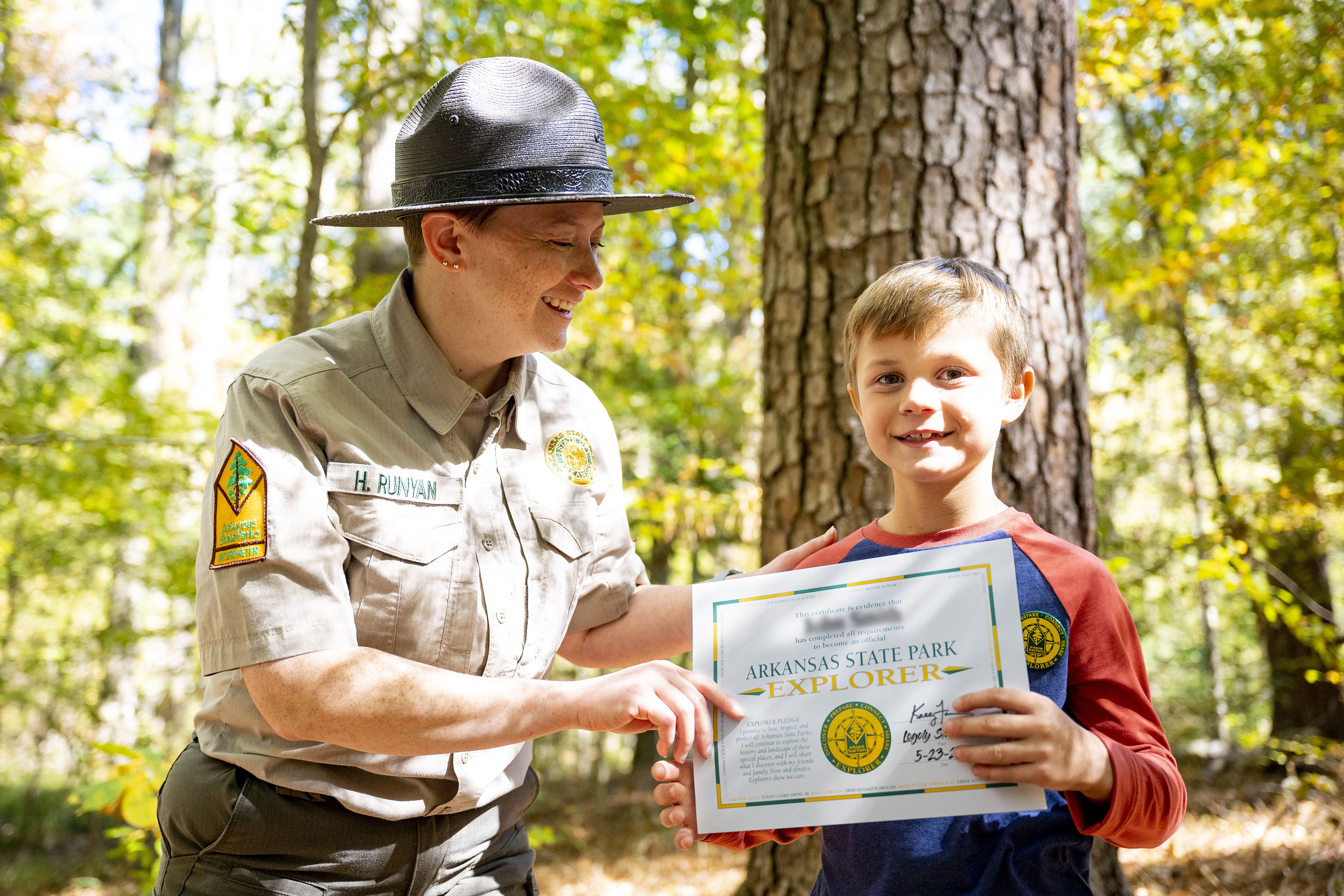 An Arkansas State Parks interpreter smiles while looking down at a child and handing them a certificate that reads Arkansas State Park Explorer. The child looks at the camera with a smile. 