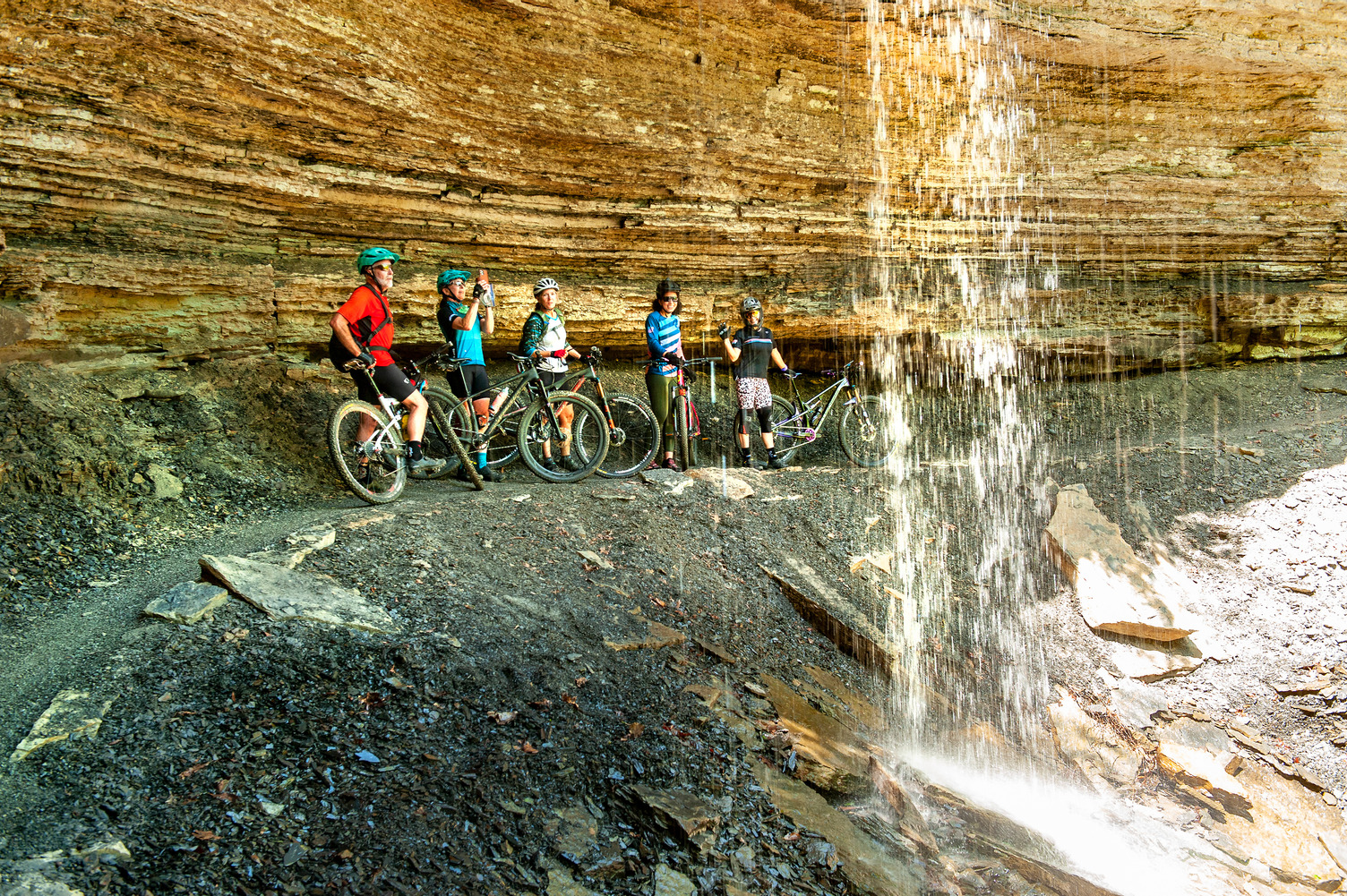 Mountain Bikers take a break under a cascading waterfall along the Devil's Den Monument Trail