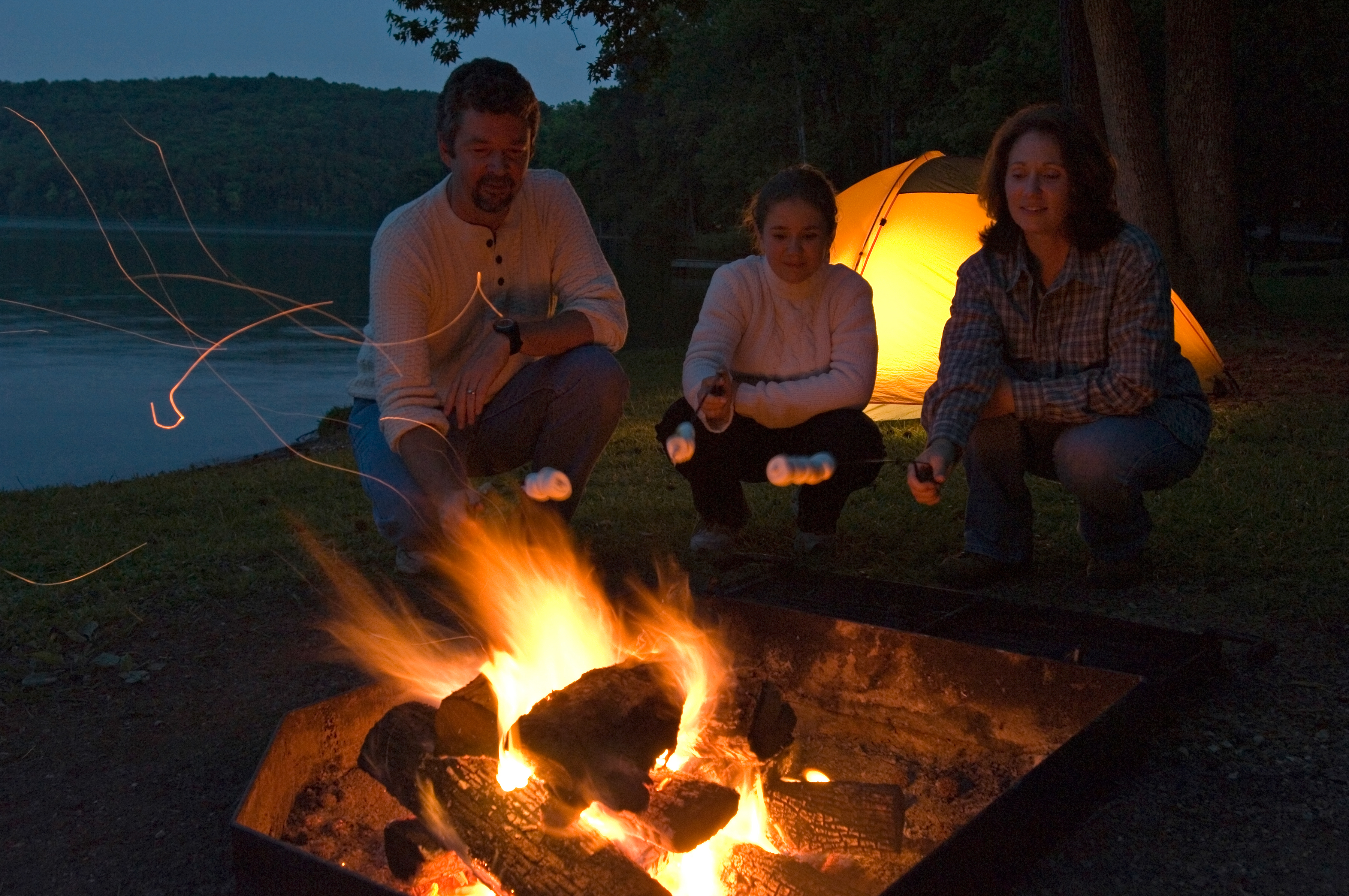 Three people squatting behind a fire roasting marshmallows with a lit up tent and a lake in the background