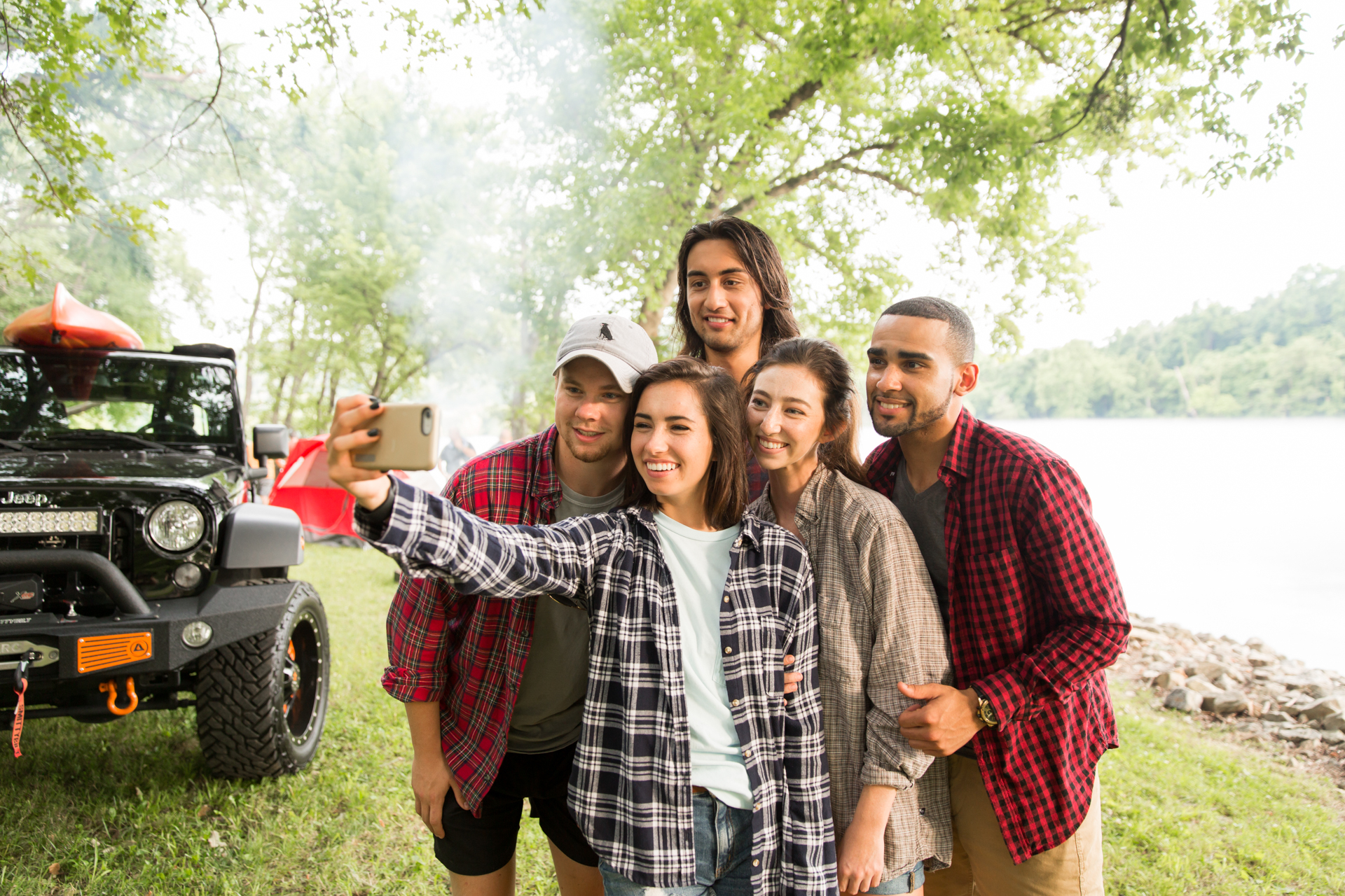 Five friends smiling and taking a selfie photo, while standing in front of a river. Their jeep is nearby with kayaks on top and a tent set up beside it.