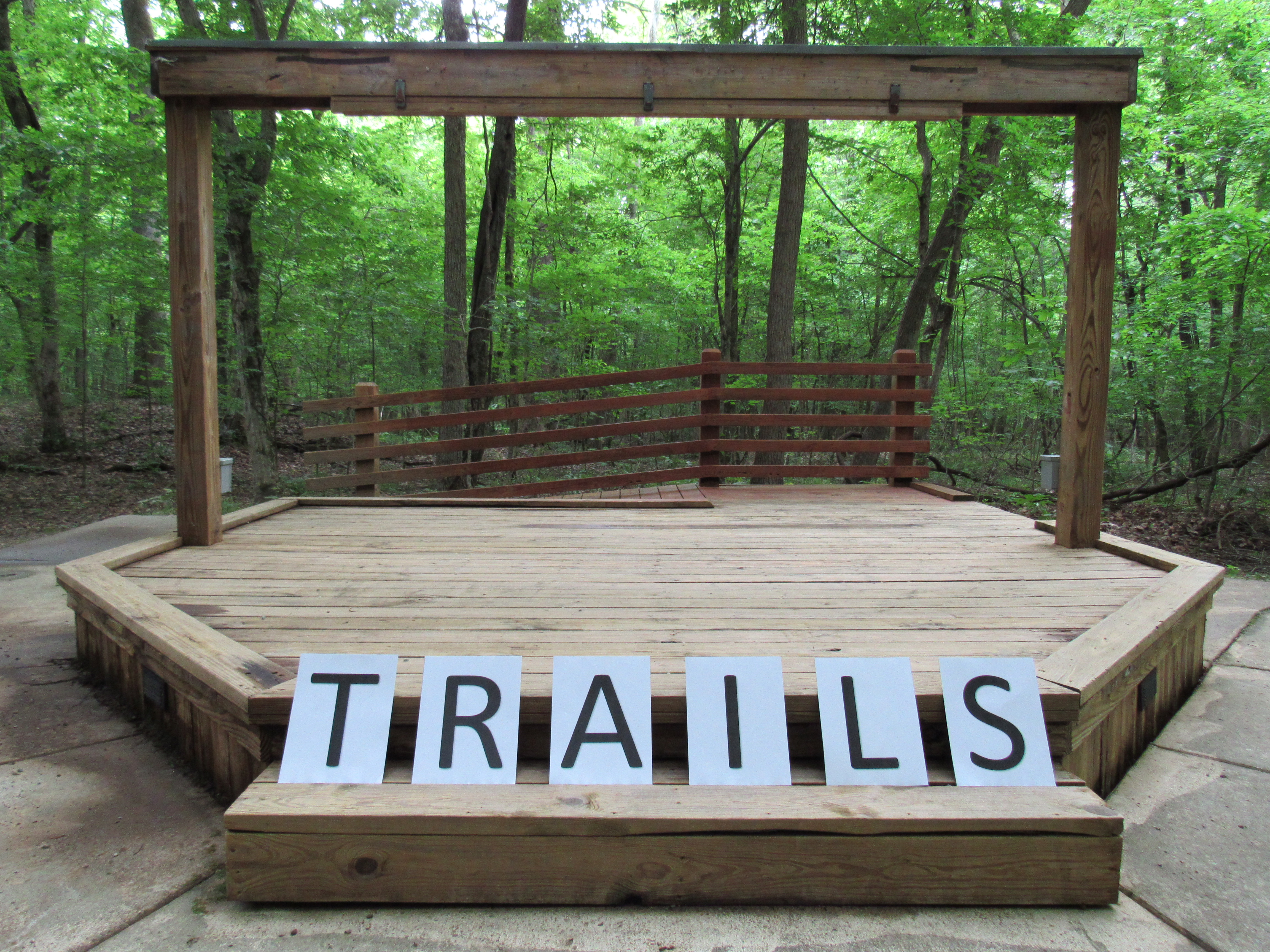 Large black letters on white paper spelling out the word TRAILS. Letters displayed on wooden amphitheater steps in a forest setting. 