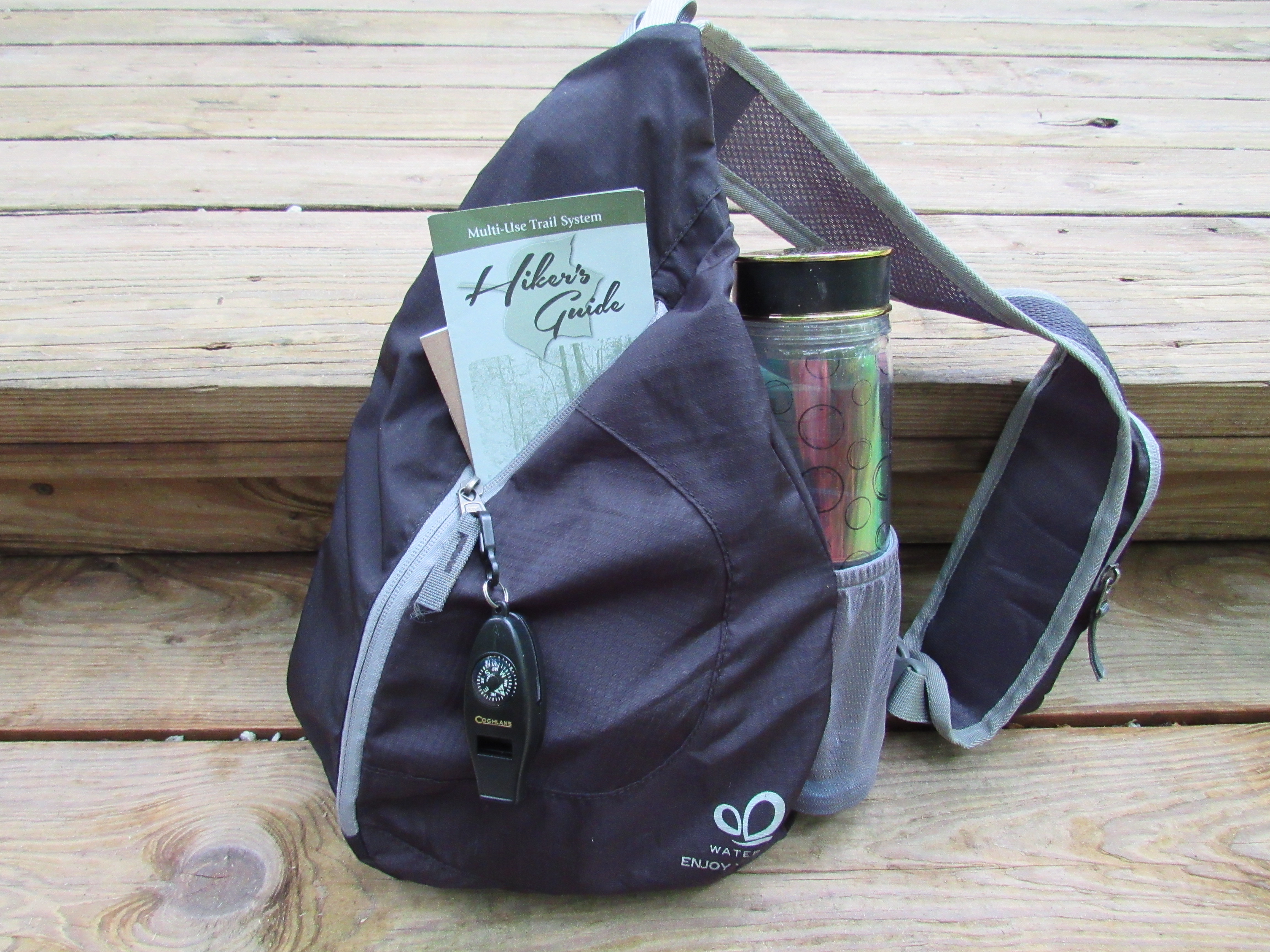 Small black backpack and a water bottle resting on wooden steps. A whistle is attached to the zipper. Sticking out of the bag is a Multi-Use Trail System Hiker’s Guide brochure. 