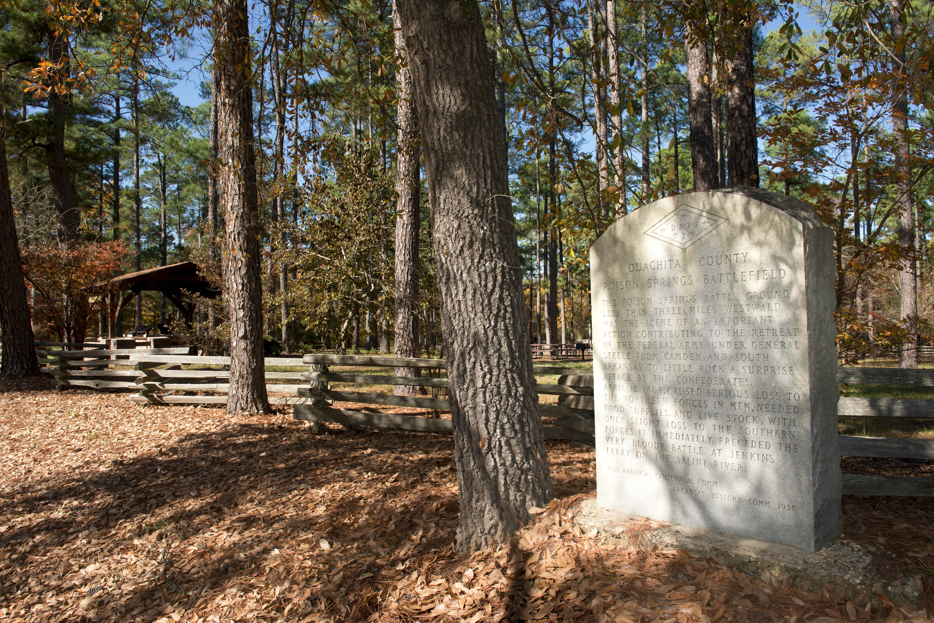 The wooded backdrop of Poison Springs with a cement marker that reads: The Poison Springs Battle Ground, less than three miles westward, was the scene of an important action contributing to the retreat of the federal army under General Steele from Camden and South Arkansas to Little Rock. A surprise attack by the confederates, April 18,1864, caused serious loss to the northern forces in men, needed food supplies, and live stock, with only slight loss to the southern forces. 