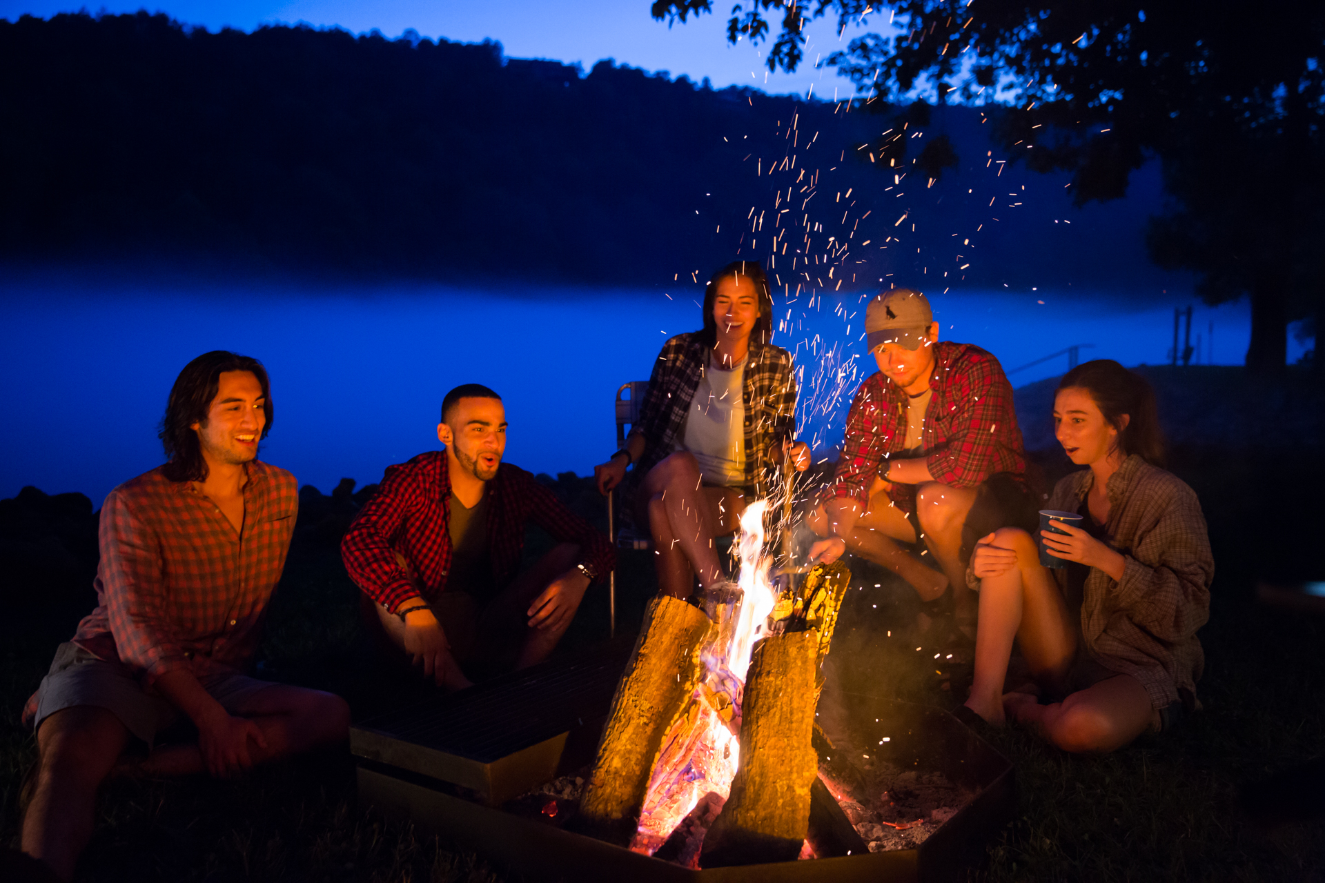 Five friends sitting around a campfire in early evening light talking and smiling; the orange flames of the fire are sparking upward into a dark blue sky and the faces of the campers are glowing in the light. A deep blue fogged-in river flows in the background.