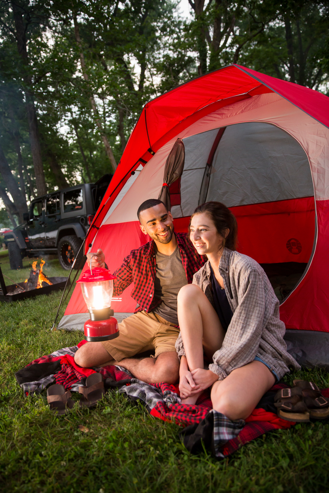 Two people sitting on a blanket outside a red tent in the forest, with a lantern for light and a campfire in the background. They are smiling and talking.