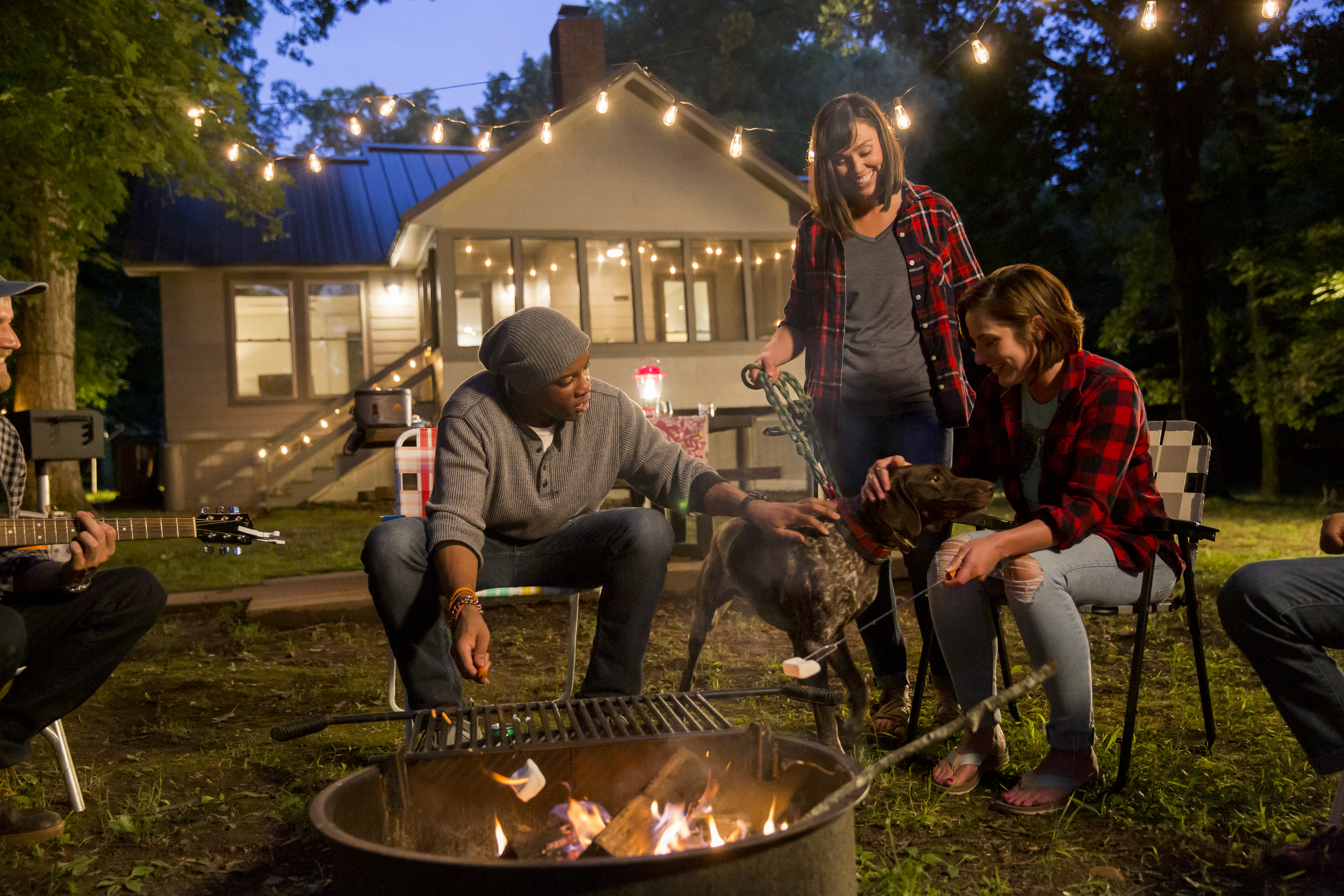 A group of people sitting around a campfire roasting marshmallows at dusk with a cabin in the background.