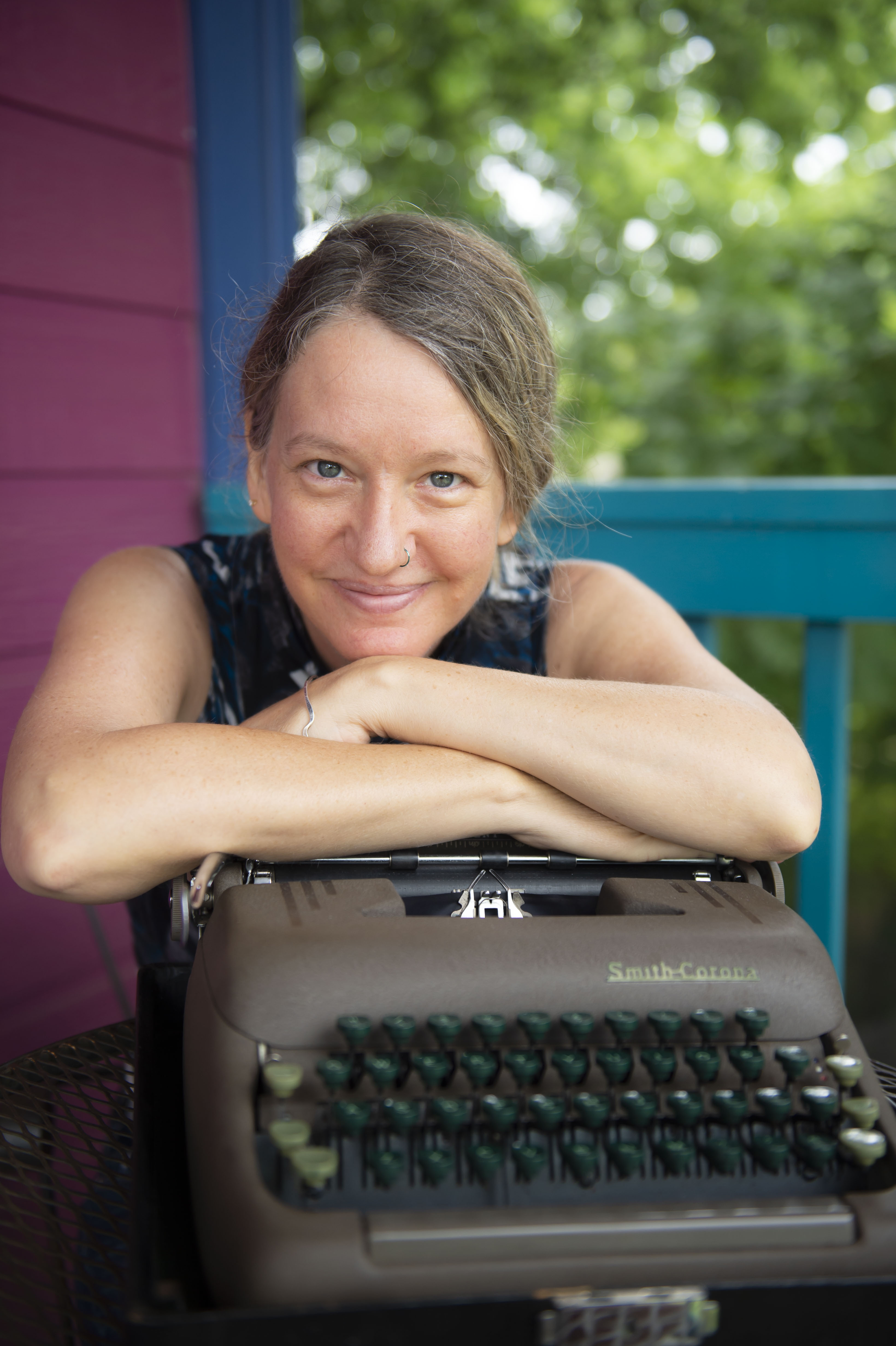 Carin Channing slightly smiling with arms folded resting on typewriter with trees in the background
