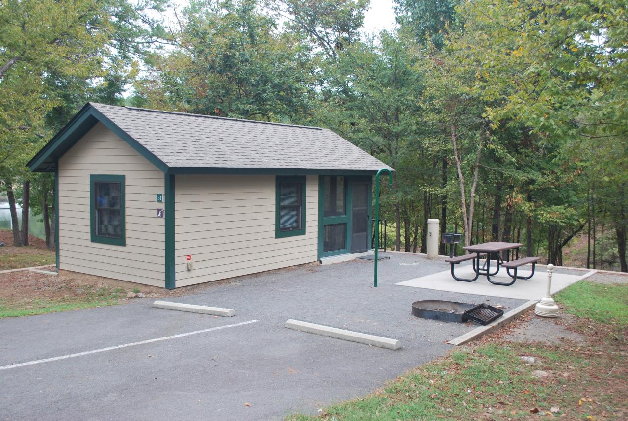 An exterior view of Camper Cabin 61 at Lake Ouachita State Park including parking spaces and an outdoor seating area with a picnic table and grill