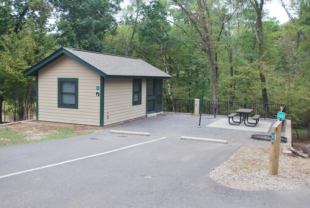 An exterior view of Camper Cabin 62 at Lake Ouachita State Park including parking spaces and an outdoor seating area with a picnic table and grill