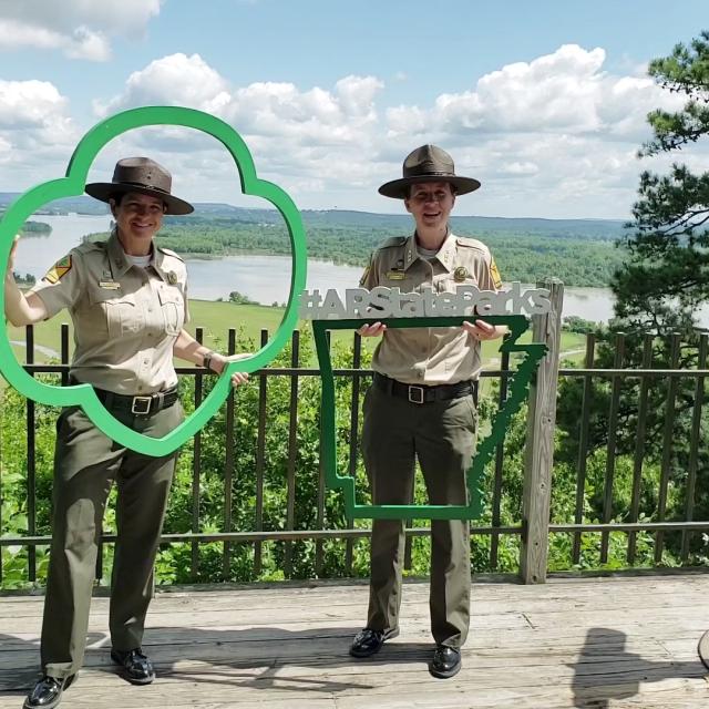 Two park rangers holding Girl Scout and Arkansas State Parks cutouts with scenic backdrop