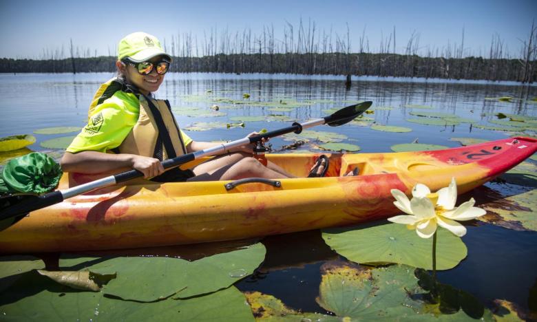 A young woman smiles at the camera while paddling along the lilypads at Cane Creek State Park