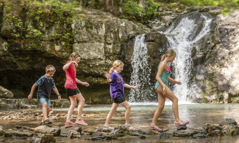 A group of children hoping along a creek with waterfalls in the background