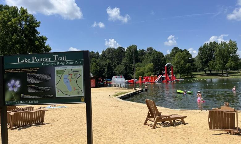 The swim beach at Crowley's Ridge State Park with Summer Splash Aqua Park in the background and Lake Ponder Trail sign in the foreground. 
