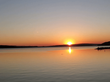 The sun sets over a wide area of Lake Dardanelle on a cloudless night. A wooden pier and rock jetty can be seen on the right, as well as distant hills on the horizon.