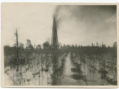 In a big, open field a wooden derrick spewing oil that covers a large flooded cotton field and a cloud of oil in the air to the right of the derrick.