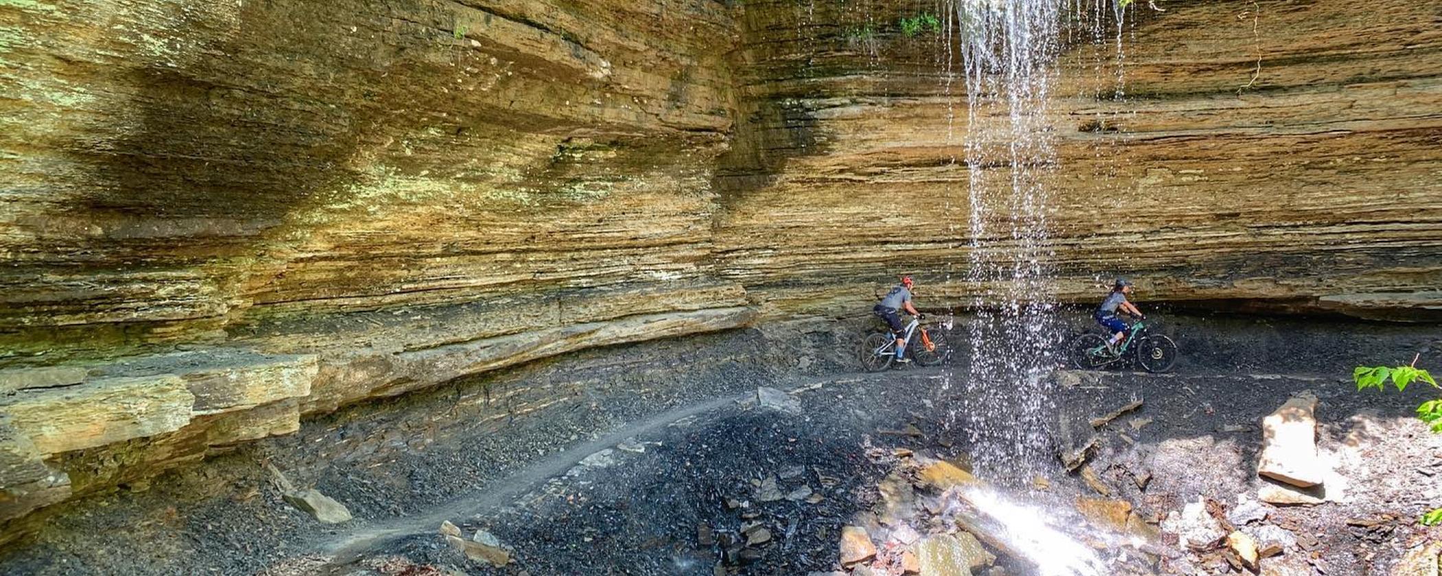 Two mountain bikers riding under a waterfall at Devil's Den State Park