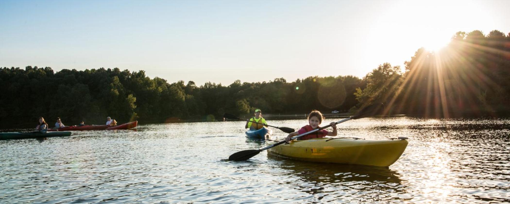 A young girl smiles at the camera in a kayak on Lake Charles with trees lining the shore and the sunset in the background