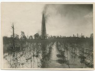 In a big, open field a wooden derrick spewing oil that covers a large flooded cotton field and a cloud of oil in the air to the right of the derrick.