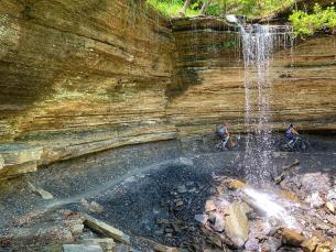 Two mountain bikers riding under a waterfall at Devil's Den State Park