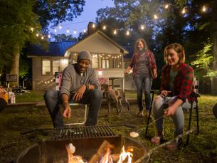 A group of people sitting around a campfire roasting marshmallows at dusk with a cabin in the background.