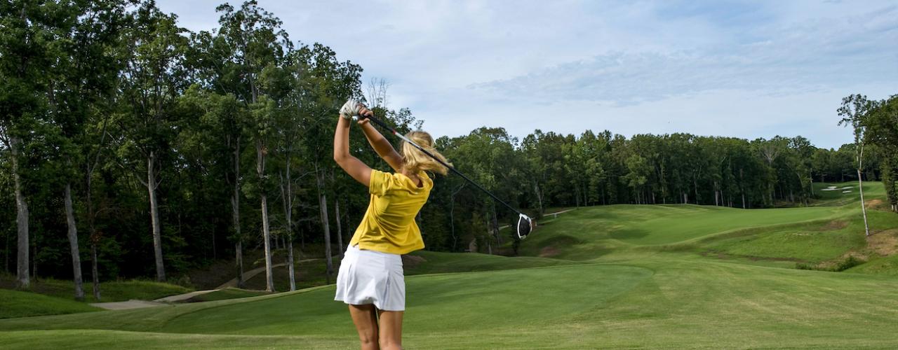 A woman, facing away from the camera, finishing her swing with a fairway lined with trees