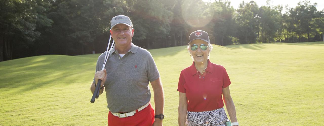 Man with a gray shirt, red shorts, gray hat with Ridges at Village Creek had holding two golf clubs against his shoulder walking towards the camera next to an older woman, smiling, with a red shirt and Red Wolves hat with a golf fairway in the background lined with trees 